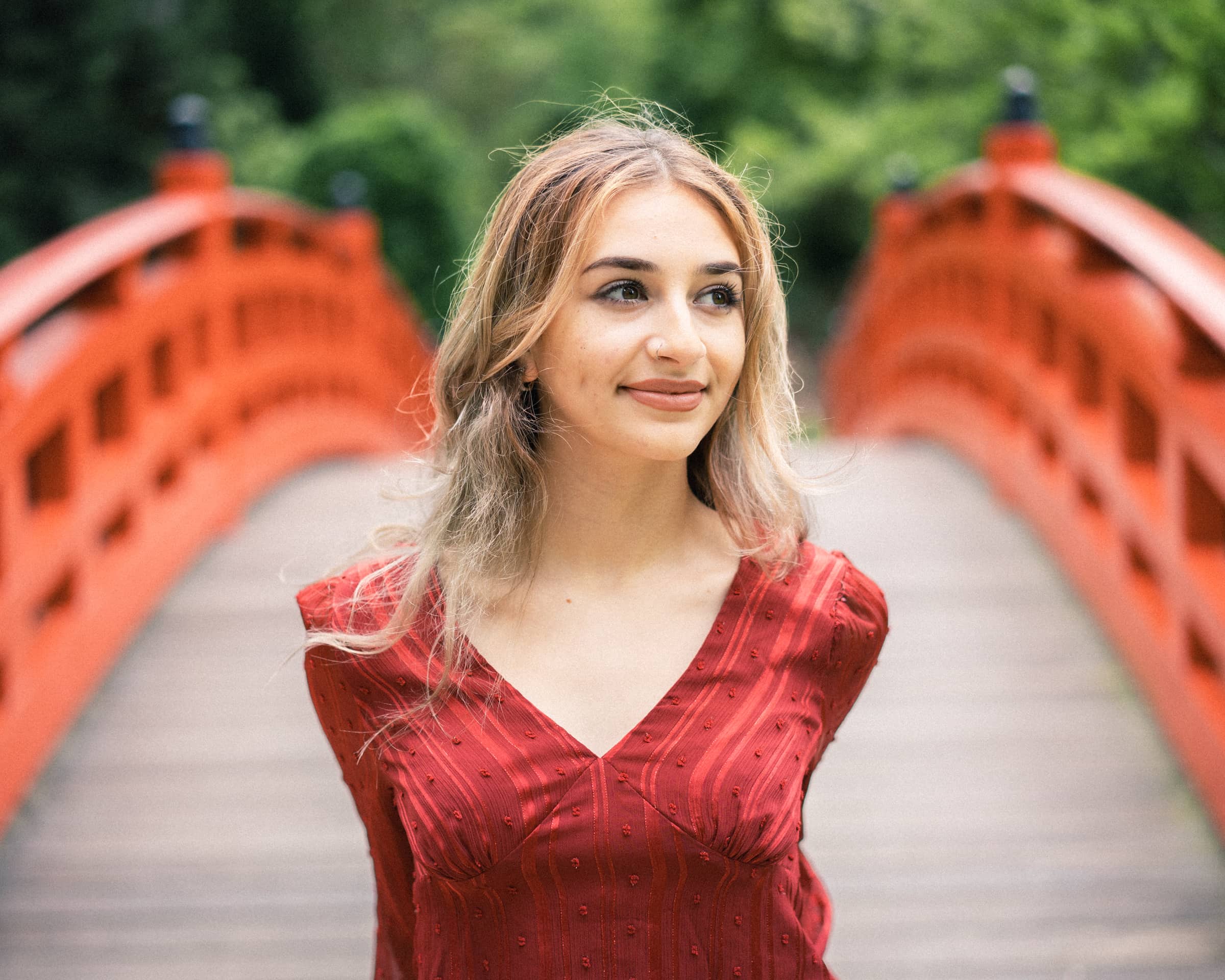 durham portrait photographers - sophia's senior session A woman with long hair stands on a wooden bridge painted red, wearing a textured red dress. Captured by a Durham portrait photographer, the scene unfolds in a lush and green backdrop, with the bridge leading into a blurred natural setting.