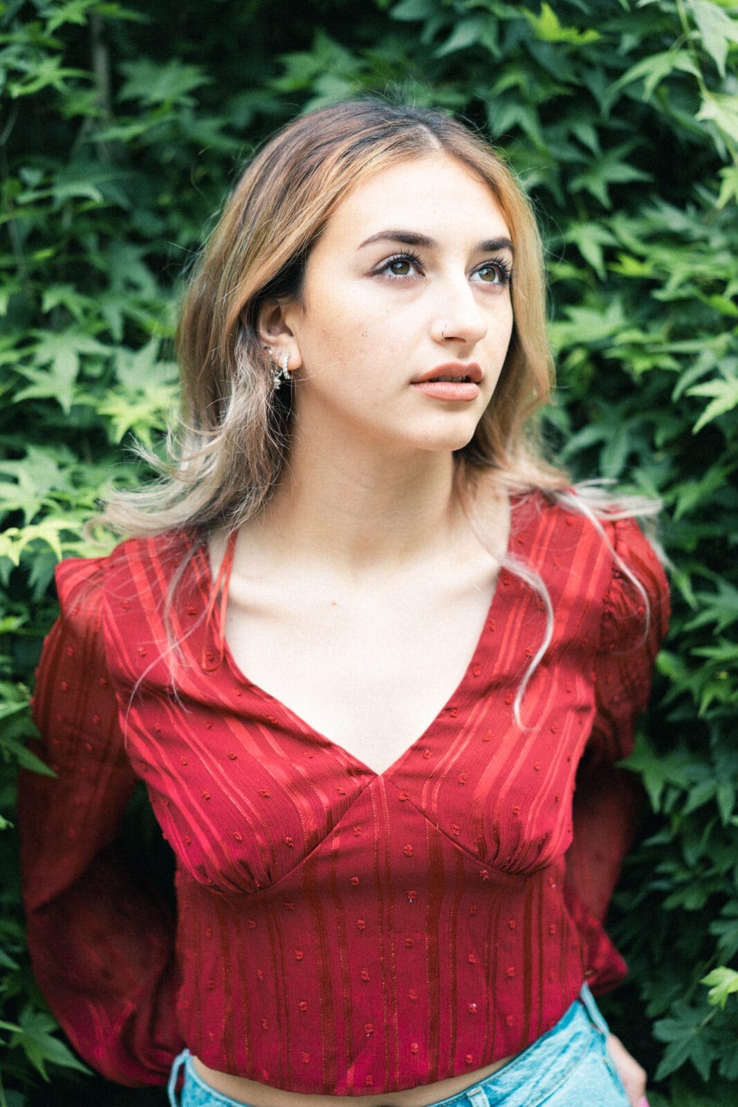 durham portrait photographers - sophia's senior session A young woman with long hair stands in front of a leafy background, captured by a Durham portrait photographer. She wears a red V-neck blouse with subtle patterns and light denim pants, looking upward with a calm expression.