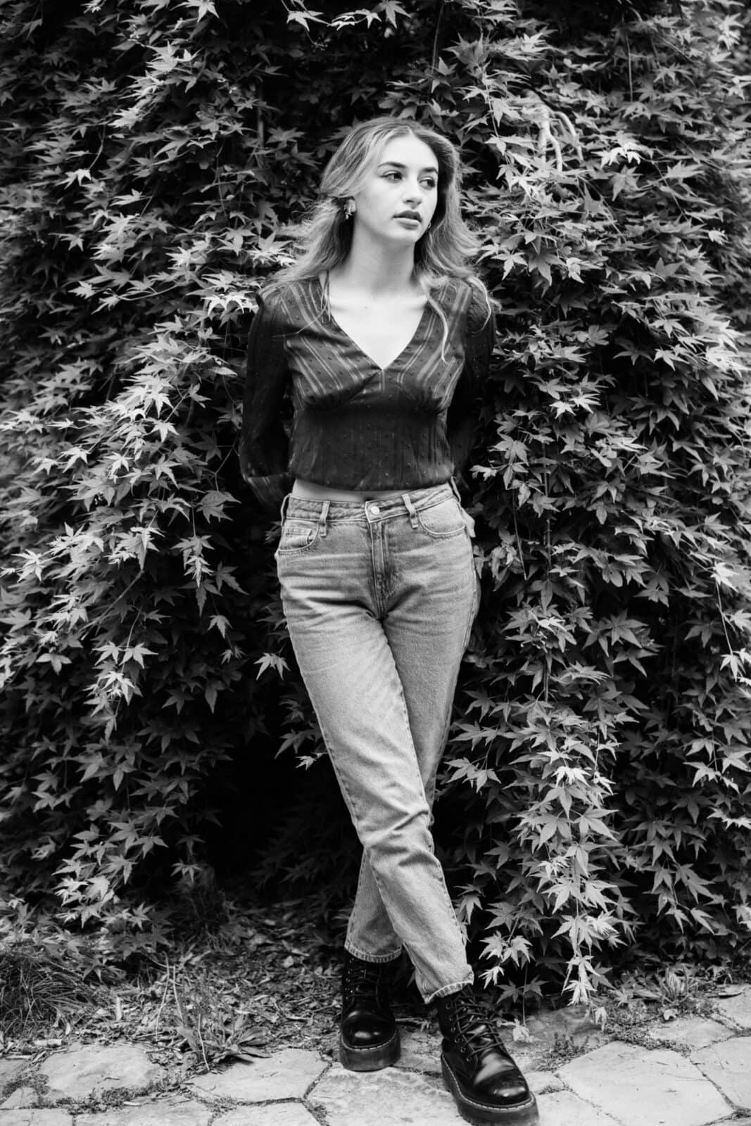 durham portrait photographers - sophia's senior session A black and white photo captures a woman standing before a lush, leafy backdrop. Her long hair flows as she wears a long-sleeved top, jeans, and boots. With hands behind her back, she gazes thoughtfully to the side—an evocative moment skillfully framed by a Durham portrait photographer.
