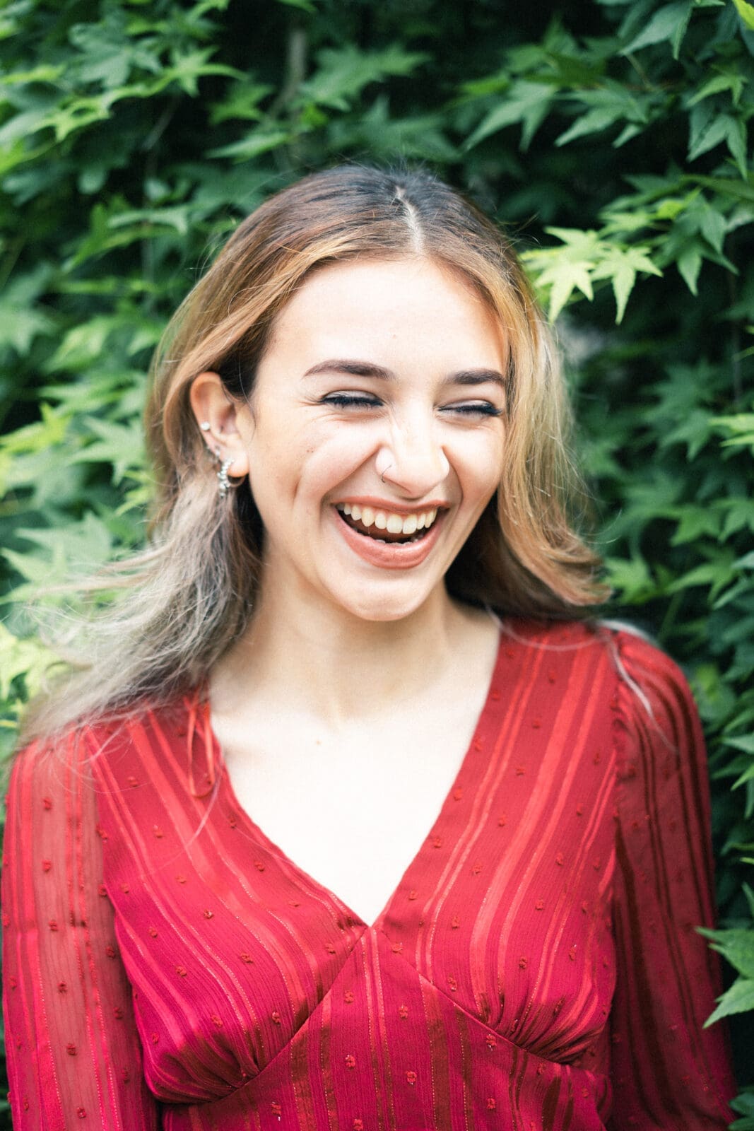 durham portrait photographers - sophia's senior session A person with long hair wearing a red blouse smiles brightly while standing in front of lush green foliage, capturing a moment reminiscent of a Durham portrait photographer's skilled eye.