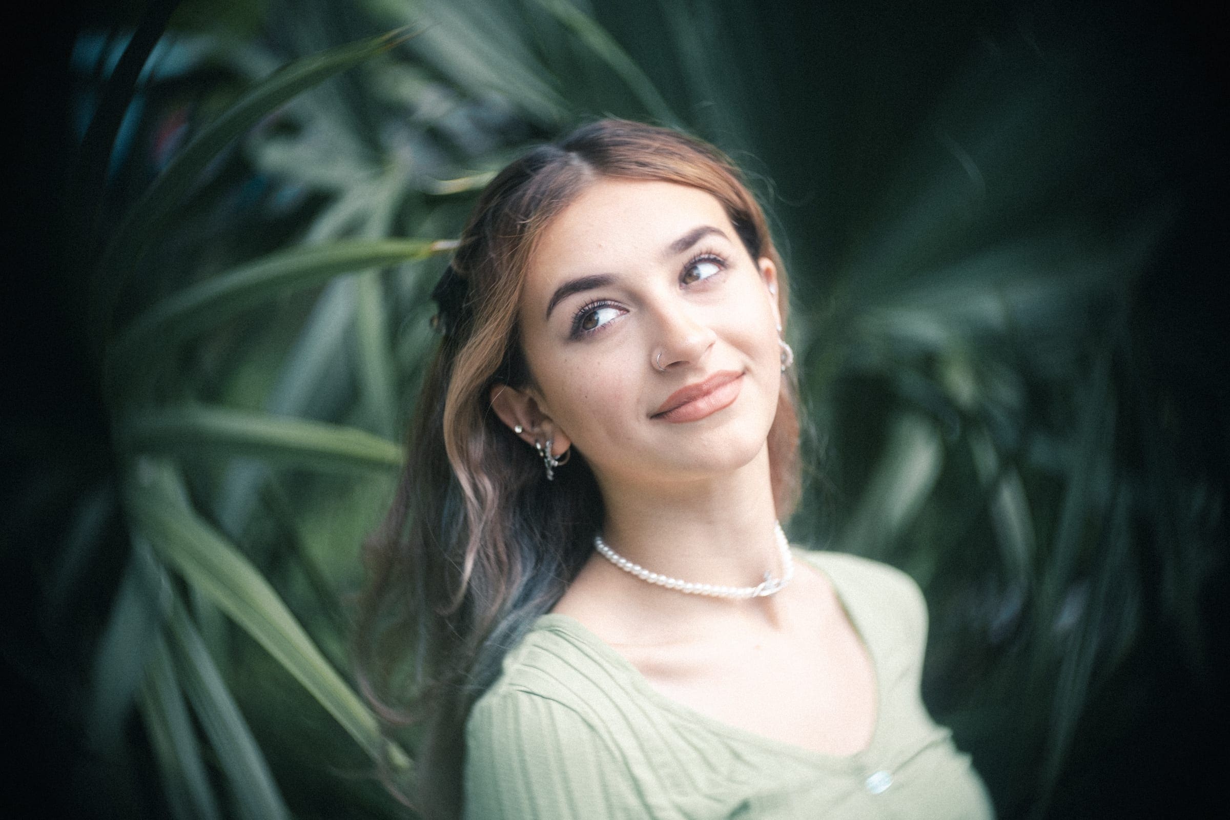 durham portrait photographers - sophia's senior session A person with long hair and a light green top stands against a backdrop of blurred greenery, captured beautifully by a Durham portrait photographer. Smiling softly, they wear pearl earrings and a necklace.