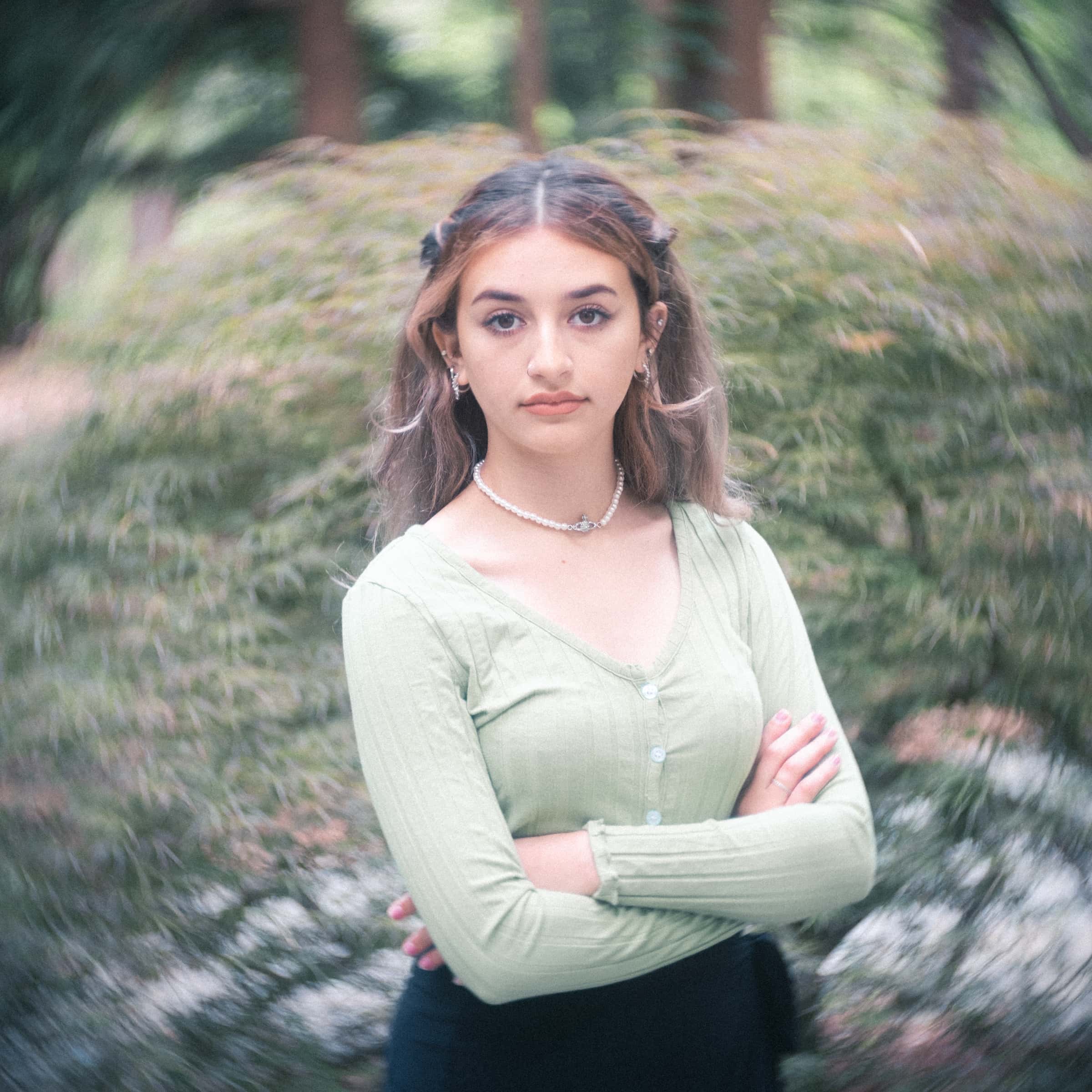 durham portrait photographers - sophia's senior session A young woman with long hair stands outdoors, arms crossed, wearing a light green long-sleeve top. Subtle jewelry complements her neutral expression. Captured by a Durham portrait photographer, she is set against a backdrop of blurred greenery.