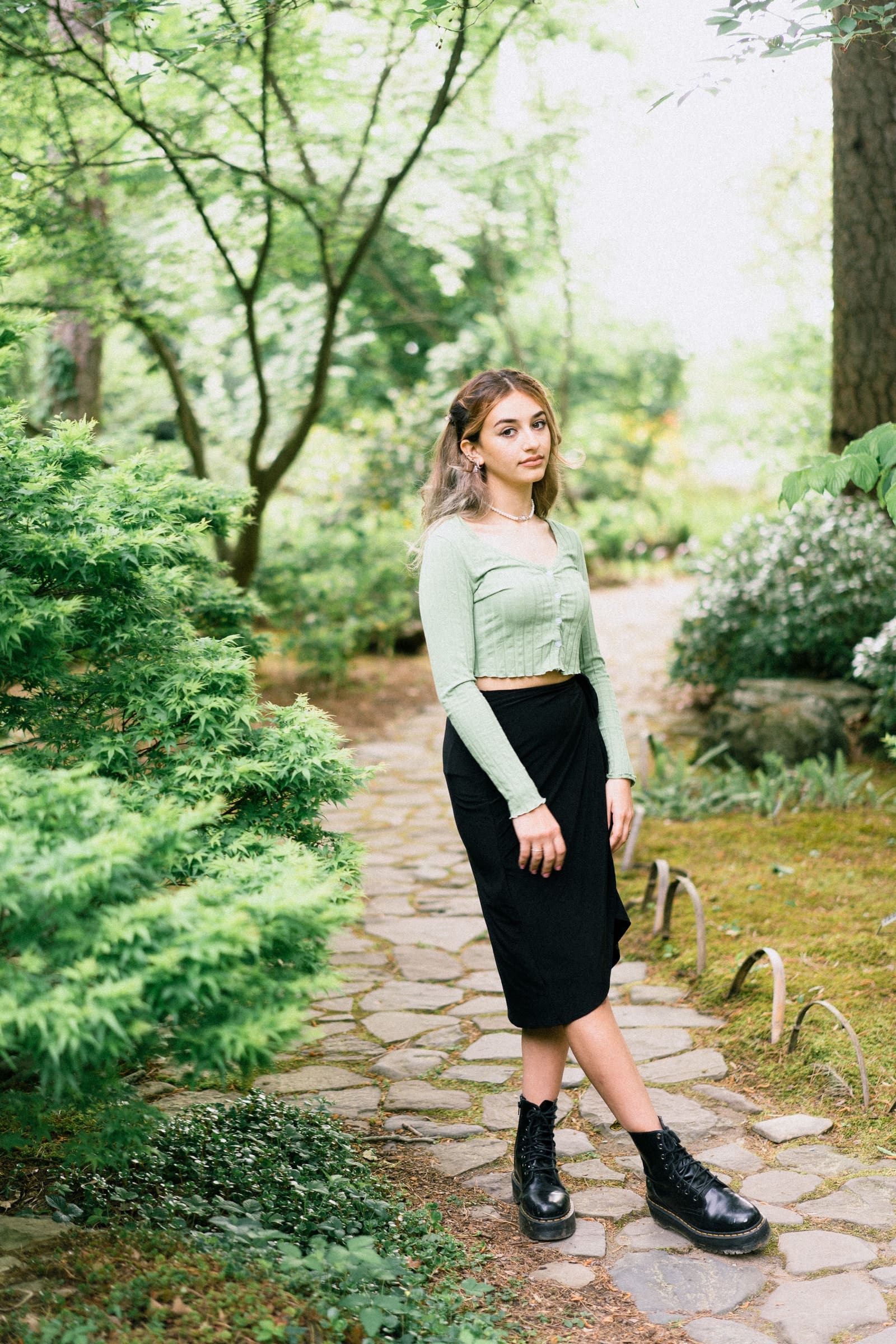 durham portrait photographers - sophia's senior session A woman in a light green long-sleeve top and black skirt stands gracefully on a stone path in a lush garden. Captured by a Durham portrait photographer, she showcases her black boots and light brown hair amidst the vibrant greenery of trees and shrubs.