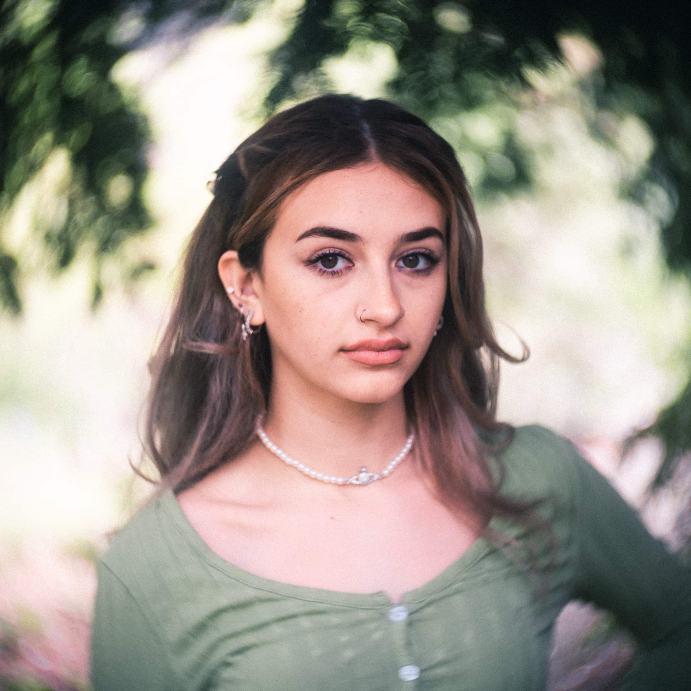 durham portrait photographers - sophia's senior session A young woman with long hair and a pearl necklace stands outdoors, captured by a Durham portrait photographer. She wears a green top and has a neutral expression, set against softly blurred foliage in the background.