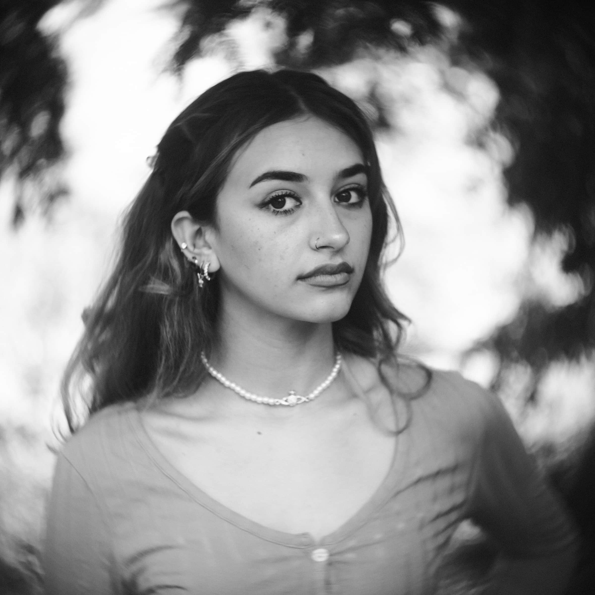 durham portrait photographers - sophia's senior session Black and white portrait by a Durham portrait photographer captures a young woman with long hair, wearing earrings and a necklace. She has a neutral expression, standing against a blurred natural background.
