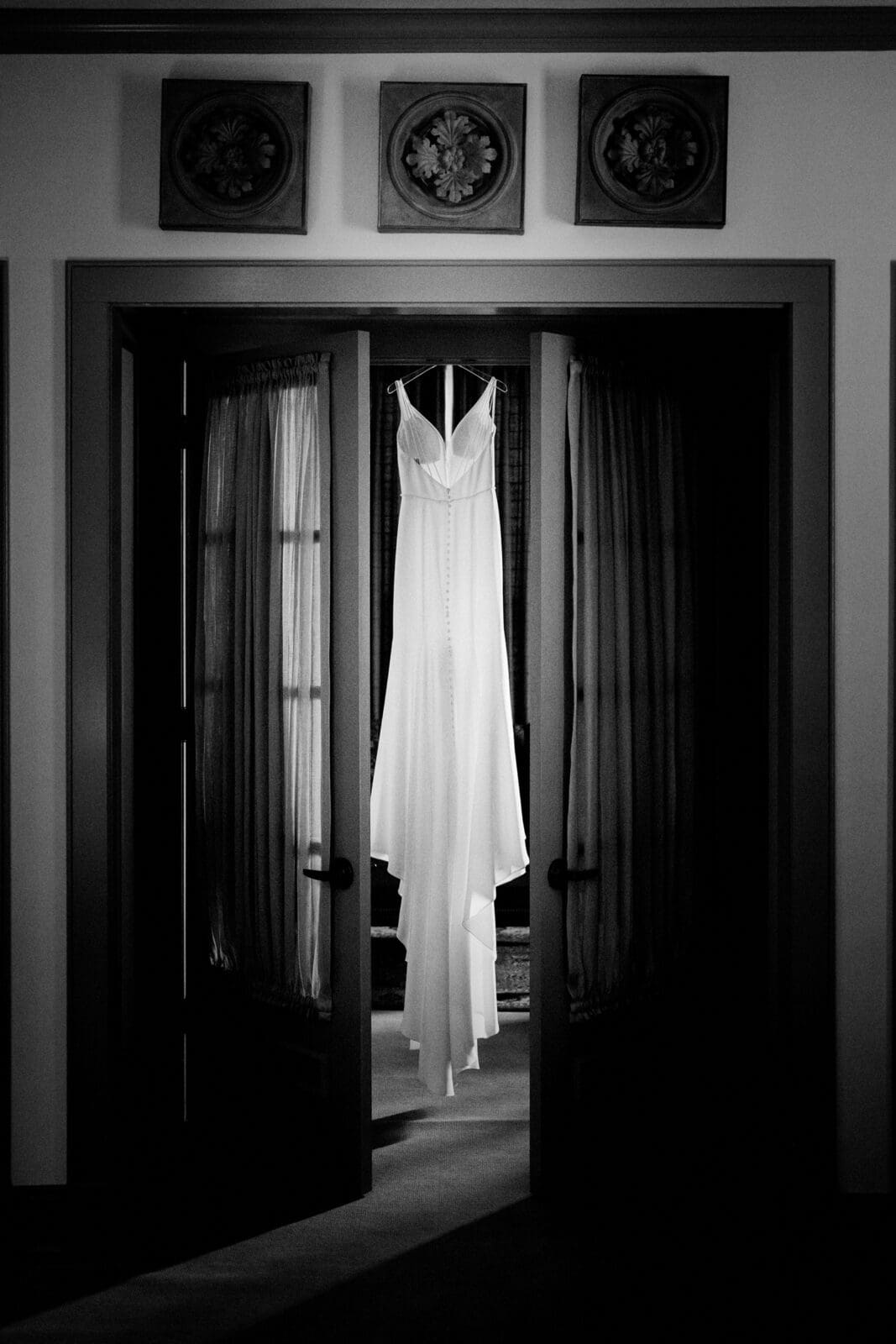 pinehurst wedding photography - lindsey & brendan A long, elegant white wedding dress hangs in the center of dark framed double doors with sheer curtains. Decorative panels adorn the wall above, creating a classic and romantic atmosphere.
