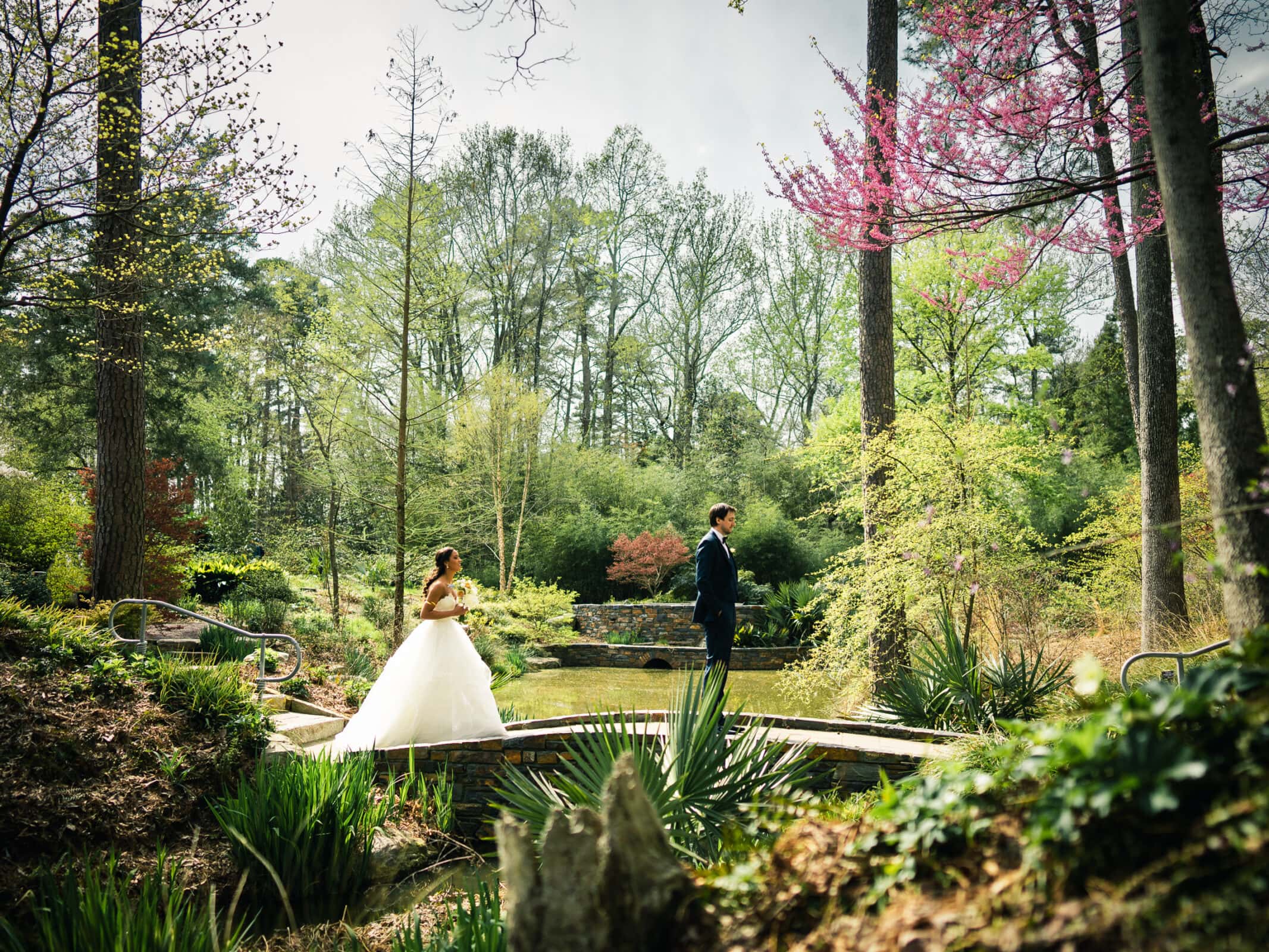 duke chapel & cookery wedding - sasha & ben A bride and groom stand on a narrow bridge surrounded by a lush garden at Duke Chapel, with trees and flowering plants. The bride wears a white gown, and the groom is in a suit, with a pond visible in the background.