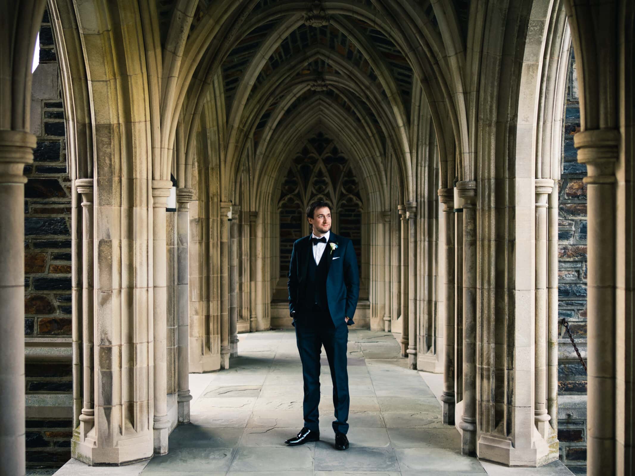 duke chapel & cookery wedding - sasha & ben A person in a dark suit stands confidently in a stone-arched hallway of Duke Chapel, with its intricate architecture and soft lighting creating a serene atmosphere.