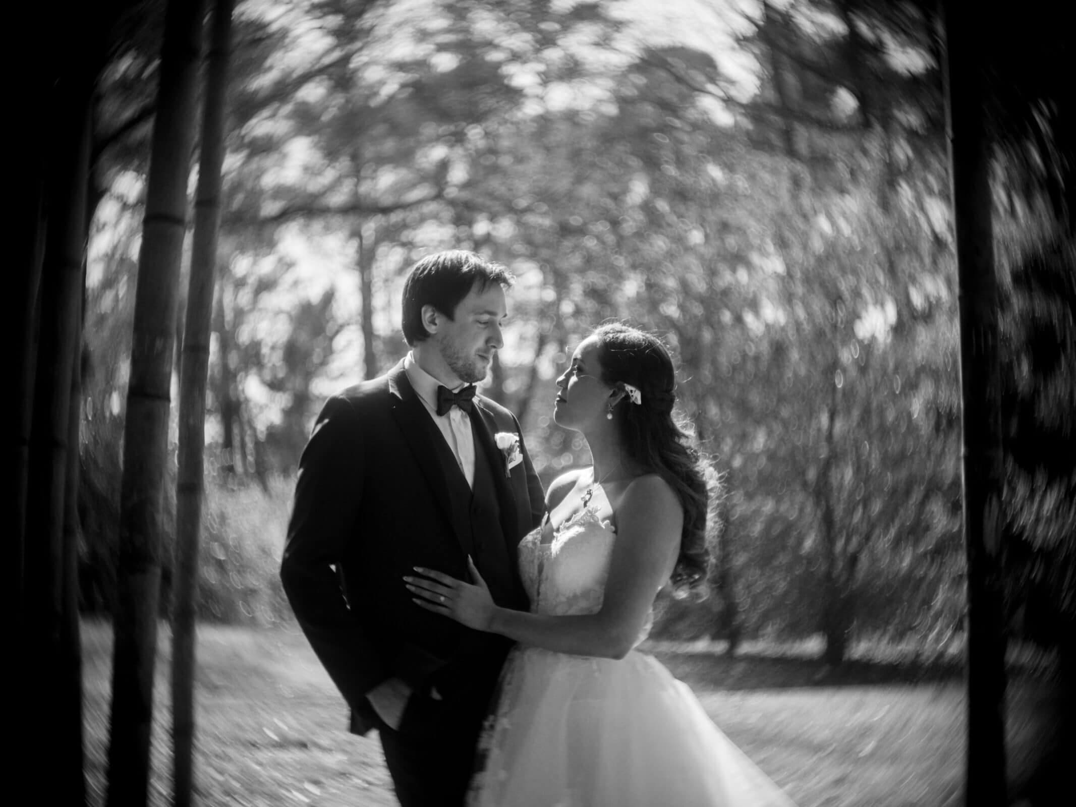 duke chapel & cookery wedding - sasha & ben A black and white photo of a couple on their wedding day, standing close in front of the iconic Duke Chapel. They gaze into each other's eyes with smiles; the groom in a suit, the bride in a gown. Blurred trees in the background create a dreamy effect.