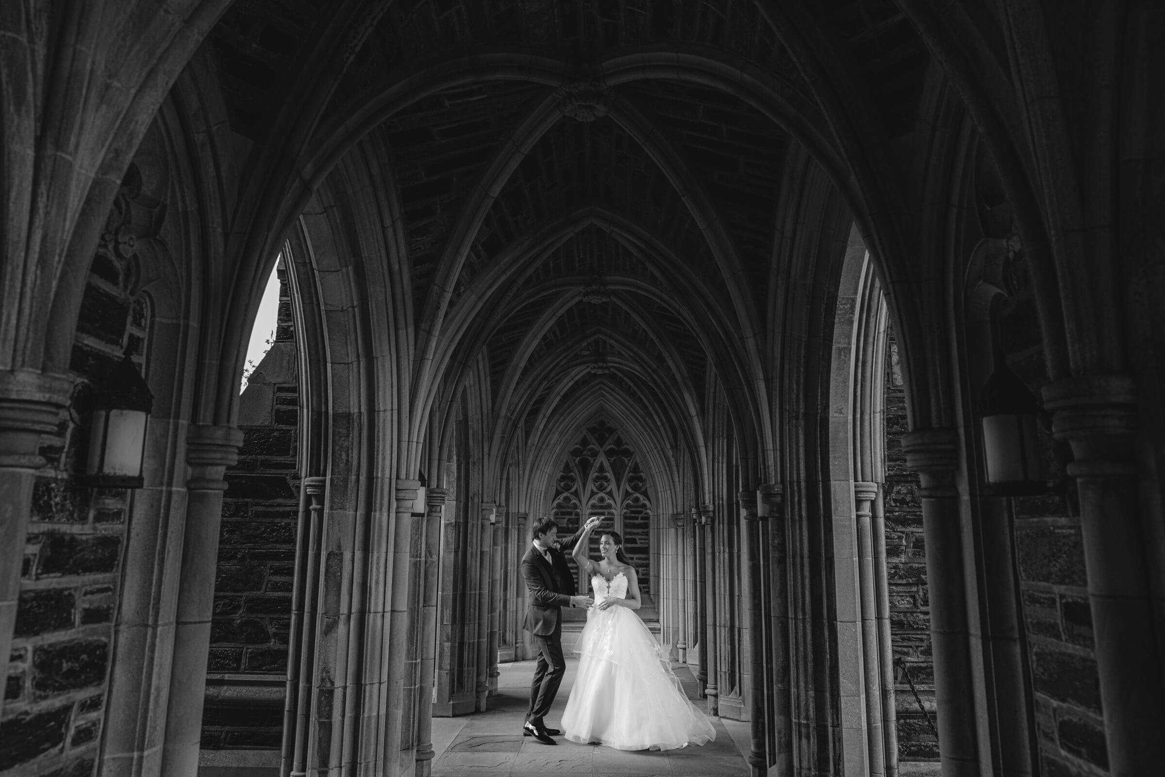 duke chapel & cookery wedding - sasha & ben A couple in wedding attire stands in the ornate, arched stone corridor of Duke Chapel. The bride wears a white dress, and the groom is in a dark suit. The setting is grand and gothic, with intricate stonework and a high-ceilinged passageway.