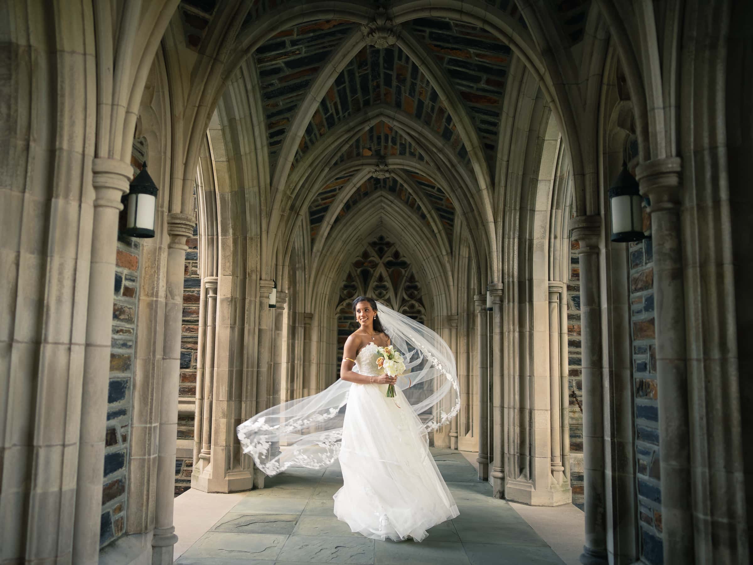 duke chapel & cookery wedding - sasha & ben A bride in a flowing white gown and veil stands joyfully in the stone archway of Duke Chapel's gothic architecture. She holds a bouquet of flowers, her dress swirling around her, creating an elegant, romantic scene.