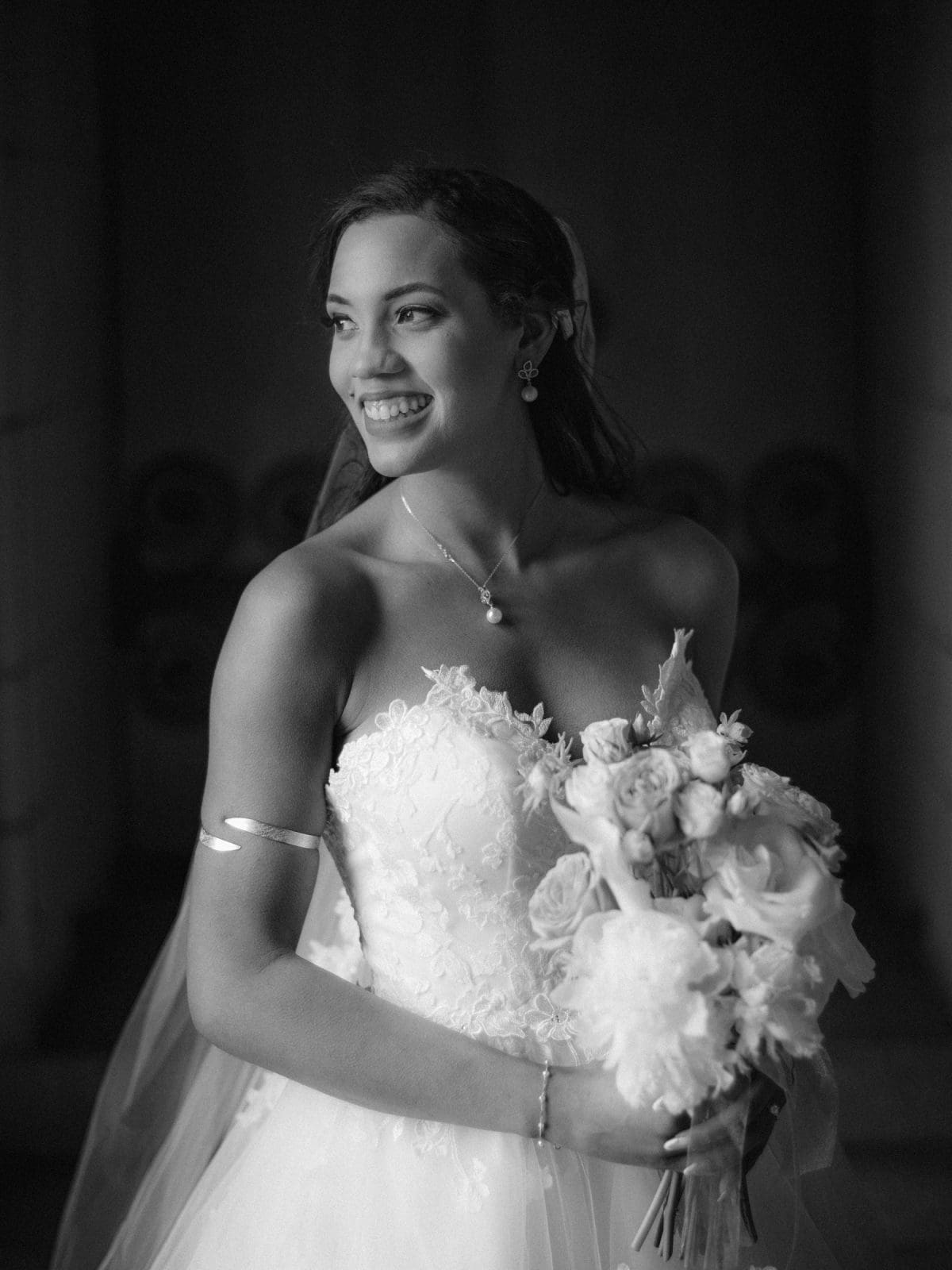 duke chapel & cookery wedding - sasha & ben A smiling bride stands indoors at Duke Chapel, holding a bouquet of flowers. She is wearing a strapless wedding gown with lace detailing and a veil. Her hair is styled with a side part, and she wears a necklace and bracelet. The image is in black and white.