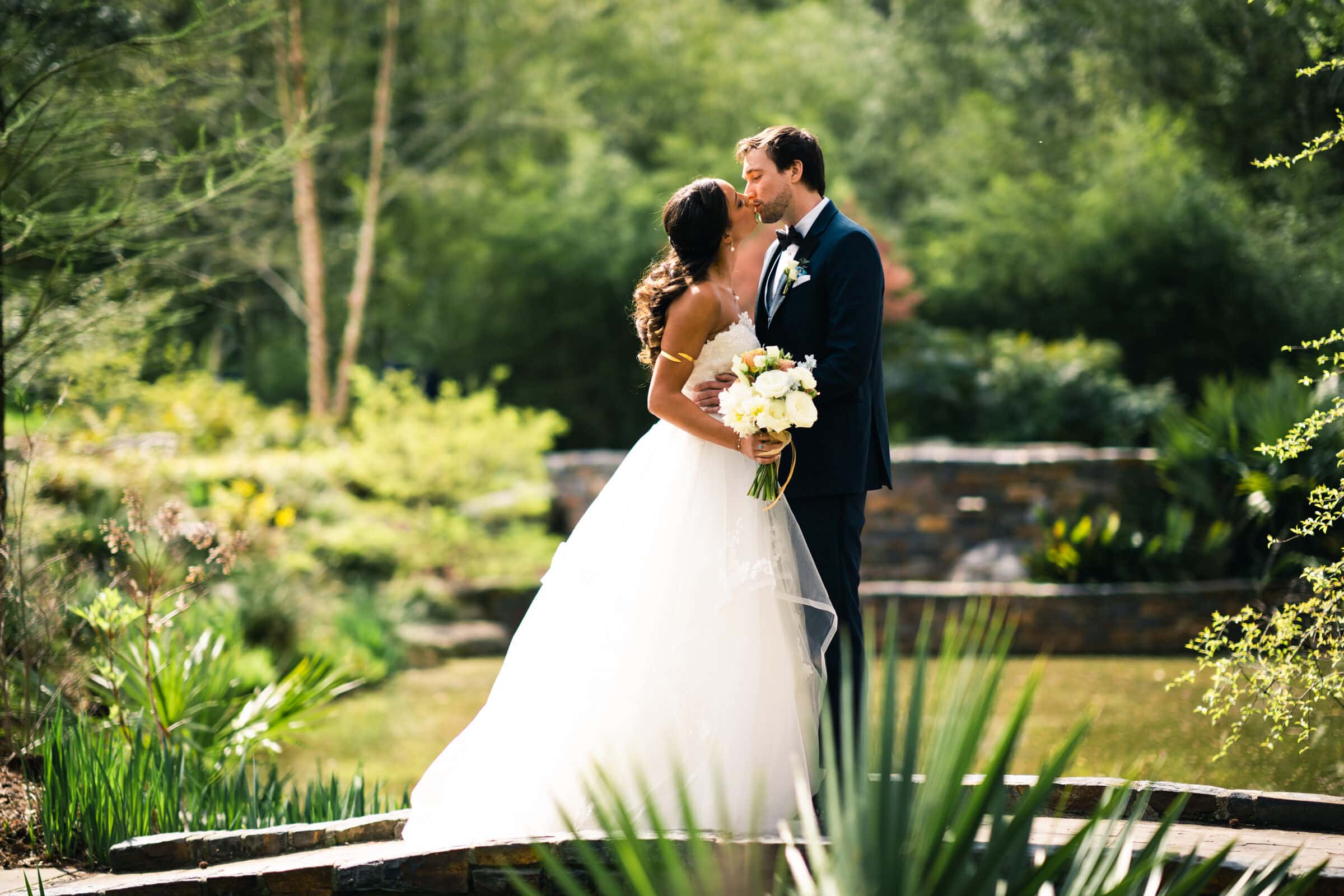 duke chapel & cookery wedding - sasha & ben A bride and groom share a kiss in a lush garden setting reminiscent of Duke Chapel. The bride, in a white gown with a bouquet of white flowers, and the groom, in a dark suit, are surrounded by green foliage and stone structures, creating a serene atmosphere.