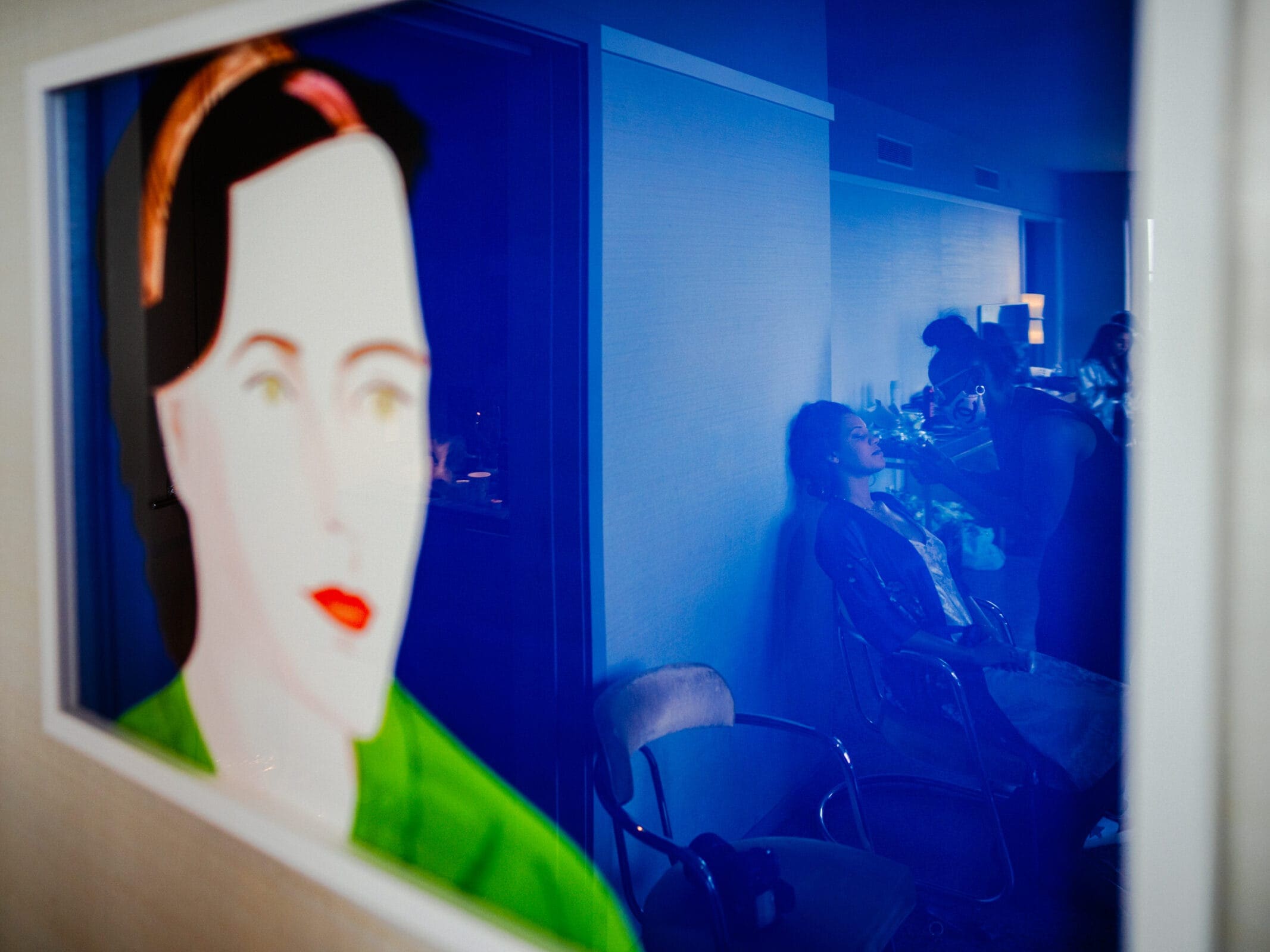 duke chapel & cookery wedding - sasha & ben In a dimly lit room with a serene blue hue, a person gets makeup applied as the soft lighting creates a relaxed atmosphere. A colorful portrait of a woman's face reflects on glass, evoking the serene beauty found within places like Duke Chapel.
