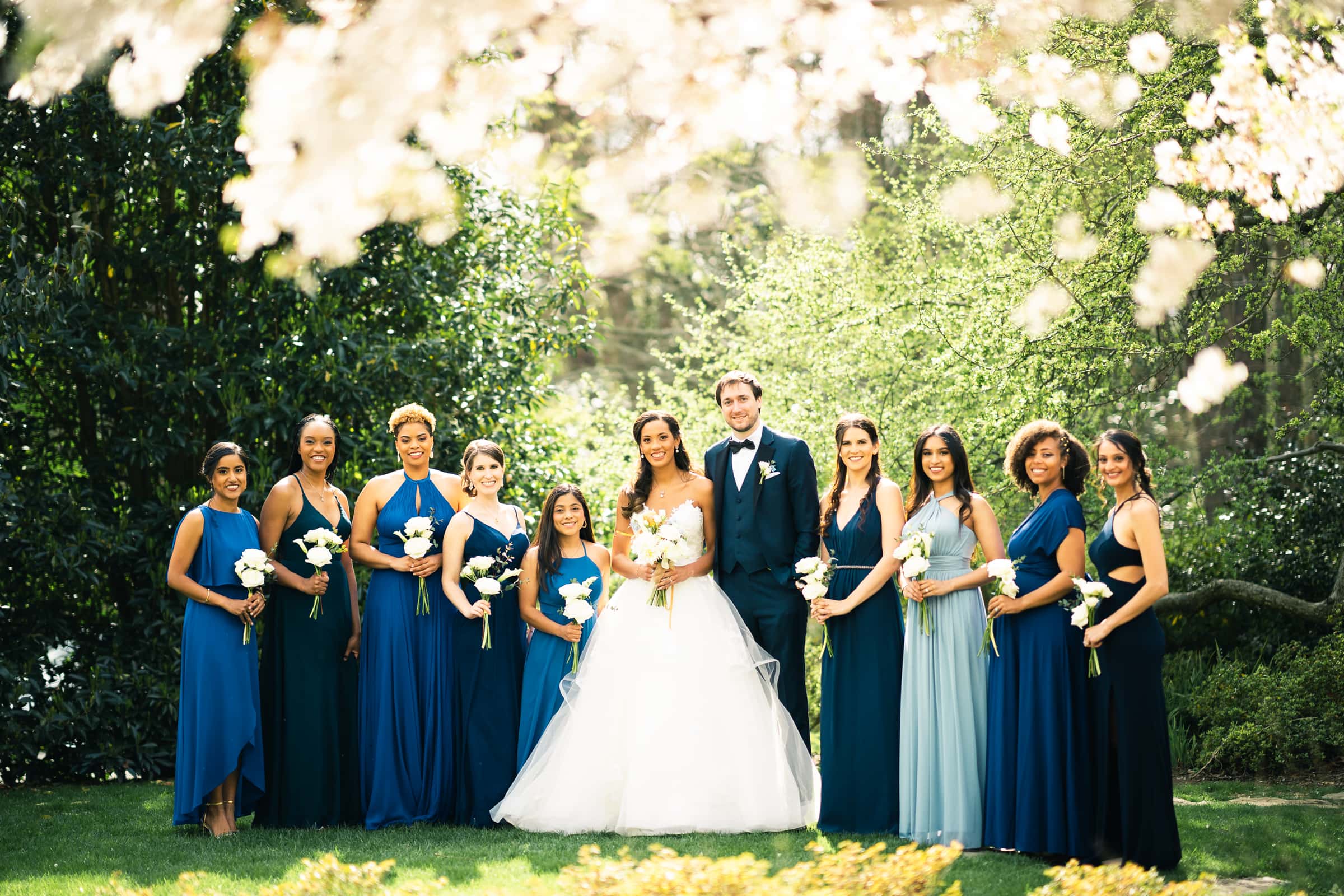 duke chapel & cookery wedding - sasha & ben A bride and groom stand together outdoors, surrounded by bridesmaids in various shades of blue dresses. They all hold white flowers, with the majestic Duke Chapel in the background, framed by lush greenery and blooming white flowers above.