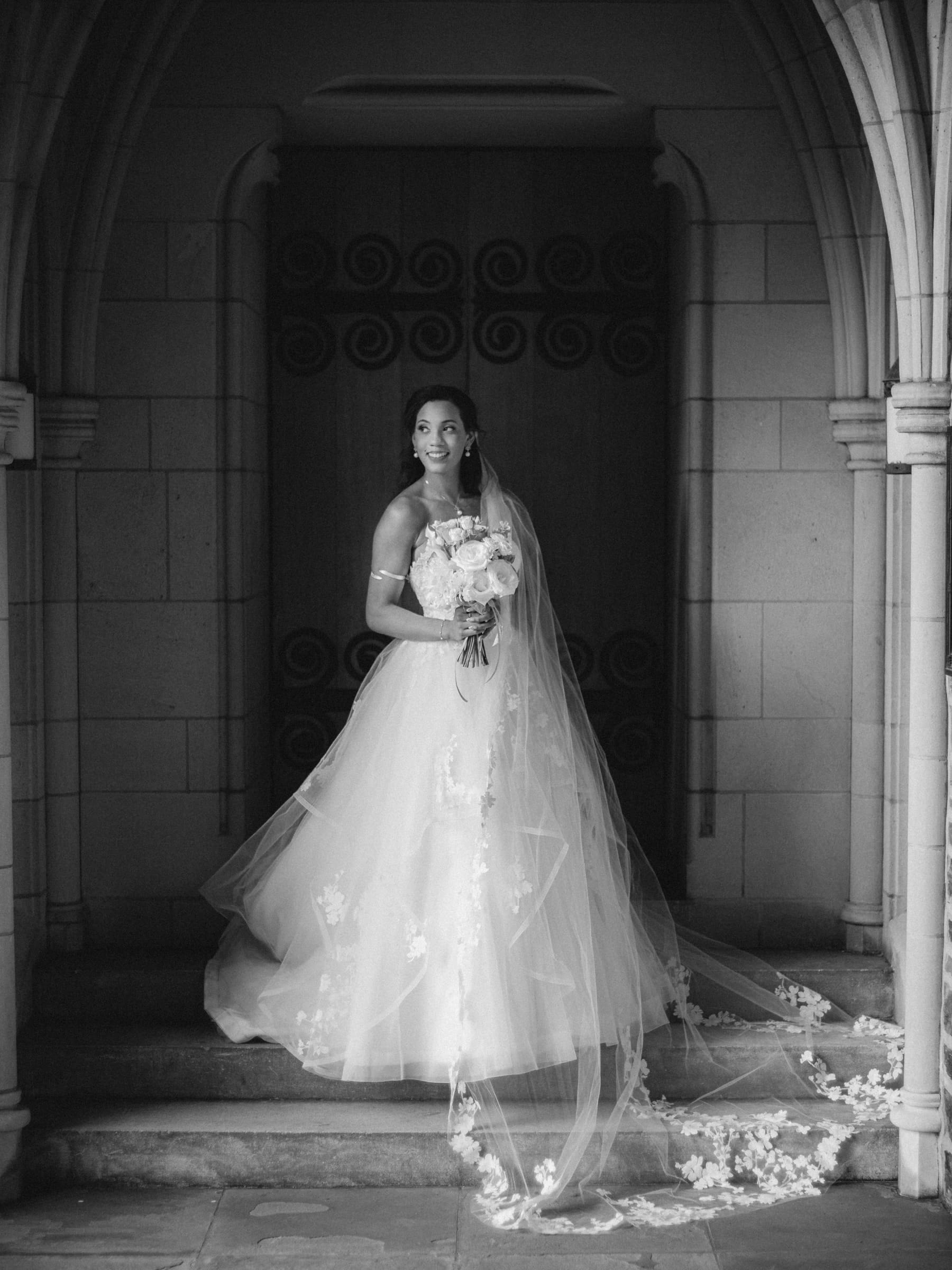 duke chapel & cookery wedding - sasha & ben A bride in a flowing, floral lace gown stands gracefully on a stone step under the arched entrance of Duke Chapel. She holds a bouquet and looks to the side with a serene expression. The black-and-white scene highlights elegant architectural details.
