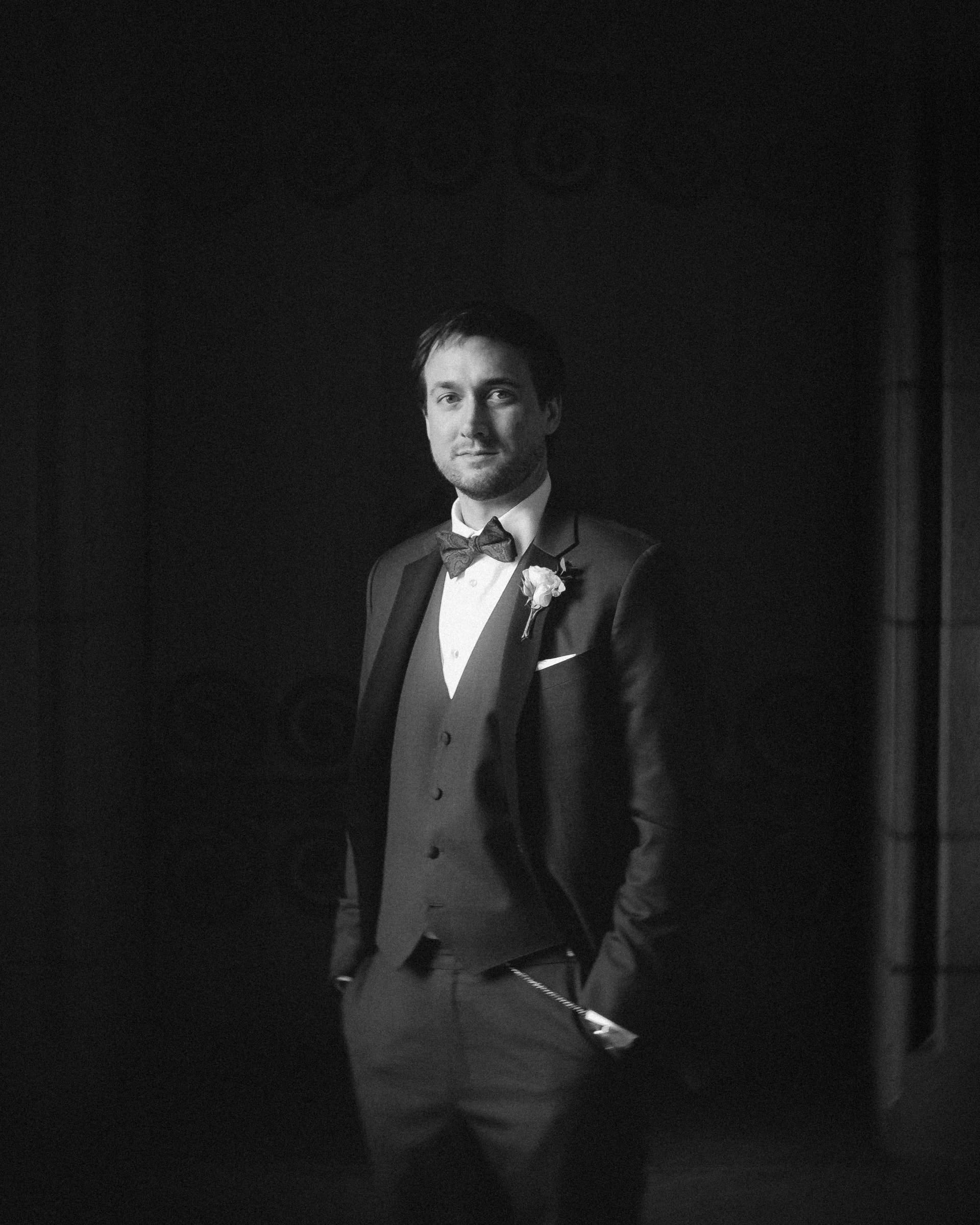 duke chapel & cookery wedding - sasha & ben A man in a formal suit with a bow tie stands with his hands in his pockets, wearing a boutonniere on his lapel. The soft, dramatic lighting mirrors the timeless elegance of Duke Chapel, creating a focused and refined atmosphere.
