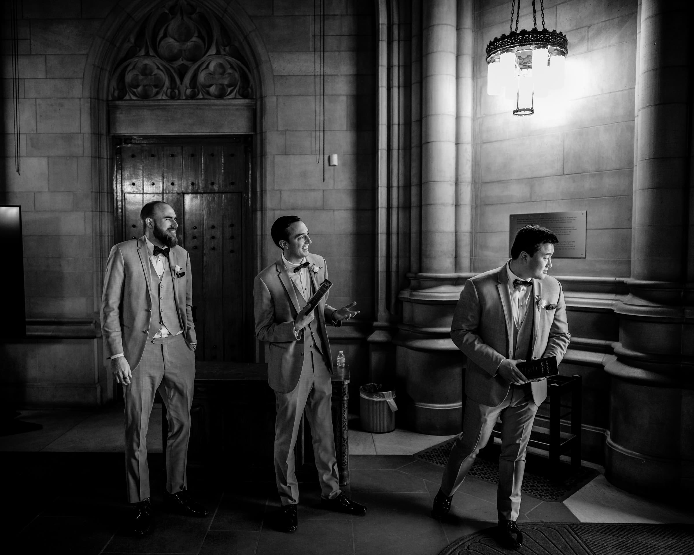 duke chapel & cookery wedding - sasha & ben Three men in formal suits stand in a dimly lit, ornate stone hallway that resembles the grandeur of a duke chapel. Two are focused on the man to their right, who is gesturing with a small box. The scene exudes a vintage, elegant atmosphere.