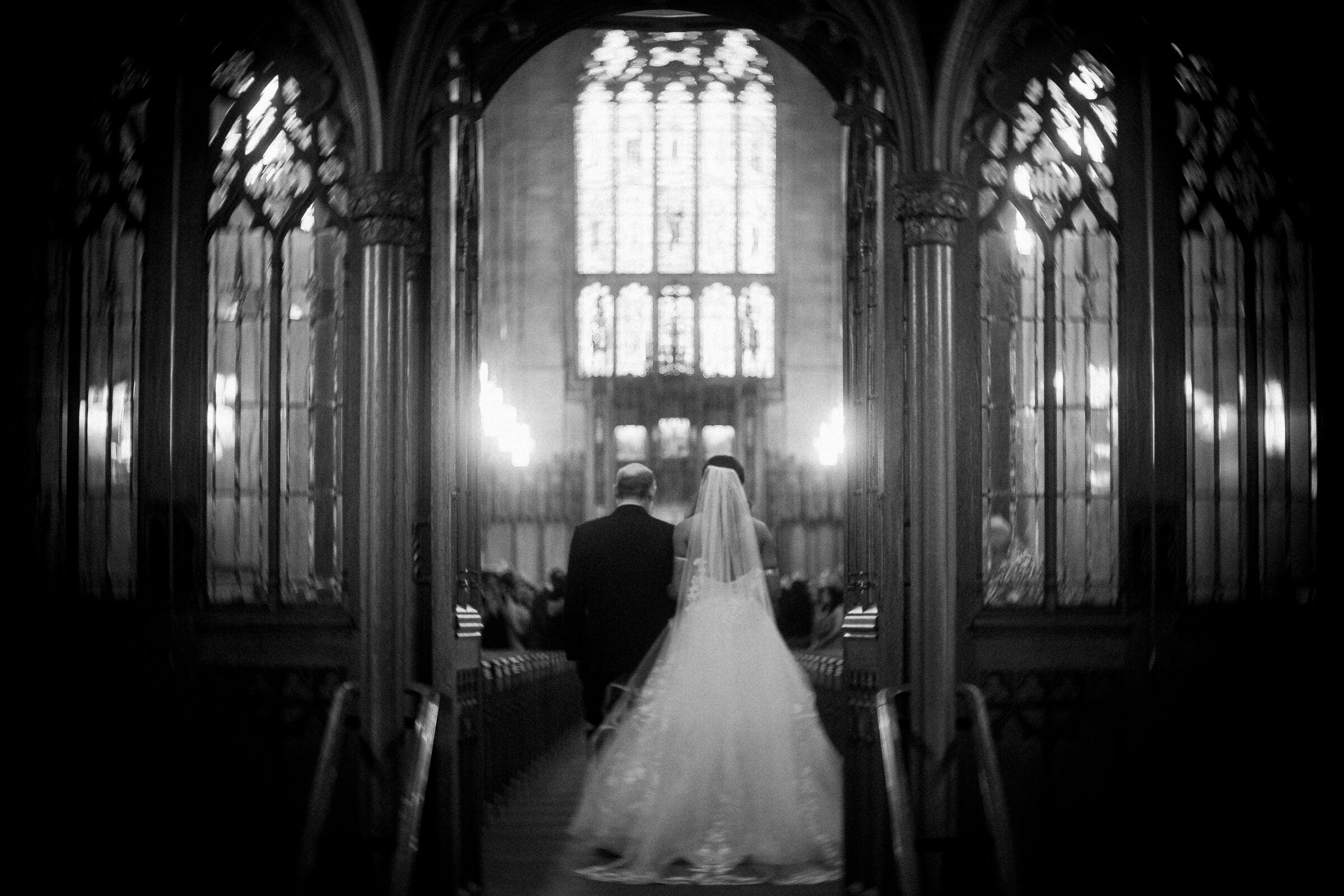 duke chapel & cookery wedding - sasha & ben A black and white image captures a bride and a man walking down an aisle in the ornate Duke Chapel. The gothic-style structure frames them with its tall, intricately designed pillars and large stained glass windows, as the bride gracefully wears a flowing gown and veil.