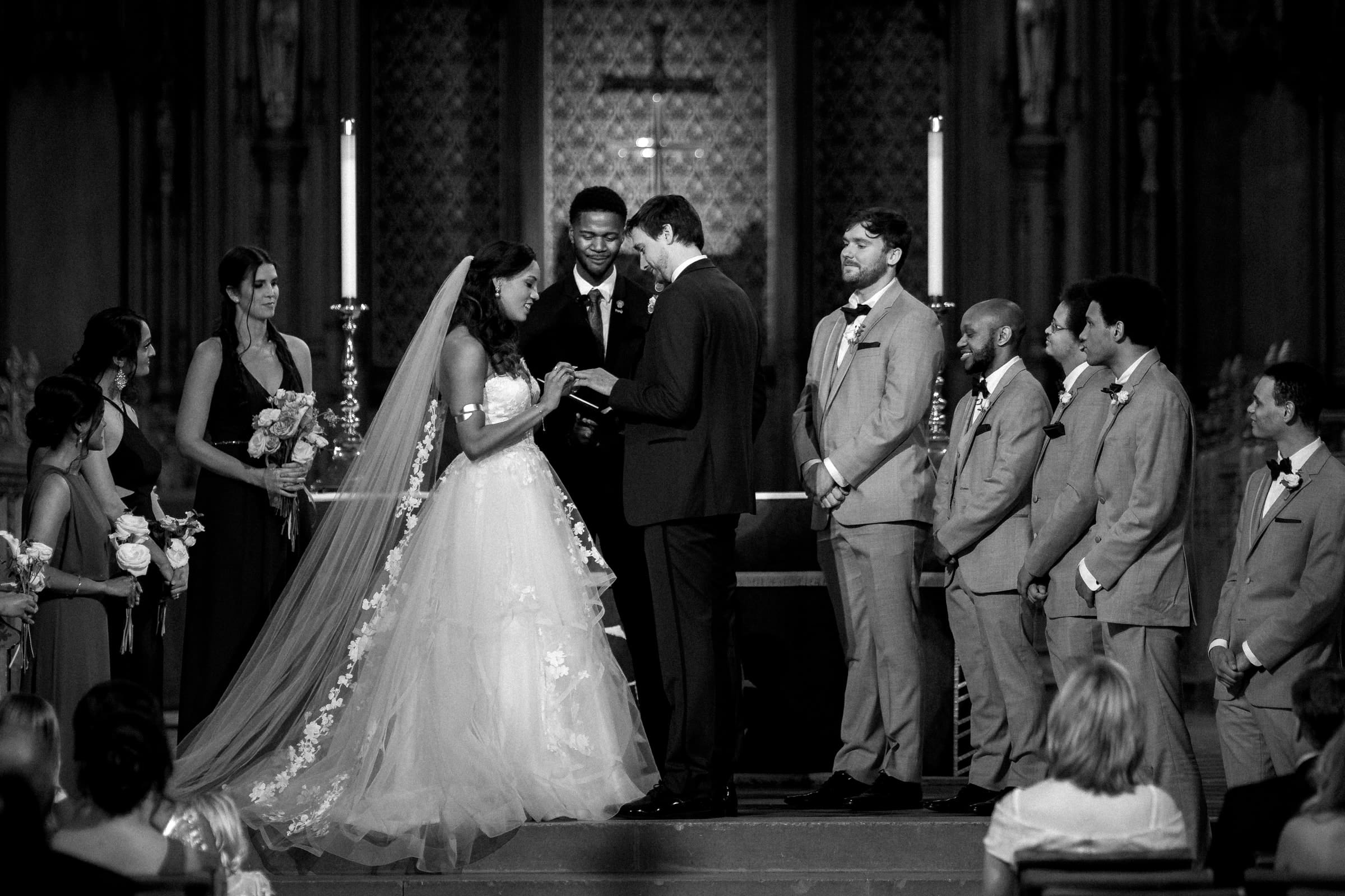 duke chapel & cookery wedding - sasha & ben A black and white photo captures a wedding ceremony at Duke Chapel. The bride and groom stand facing each other, holding hands, with the bridal party beside them—bridesmaids in dresses and groomsmen in suits—as the officiant oversees the sacred vows.