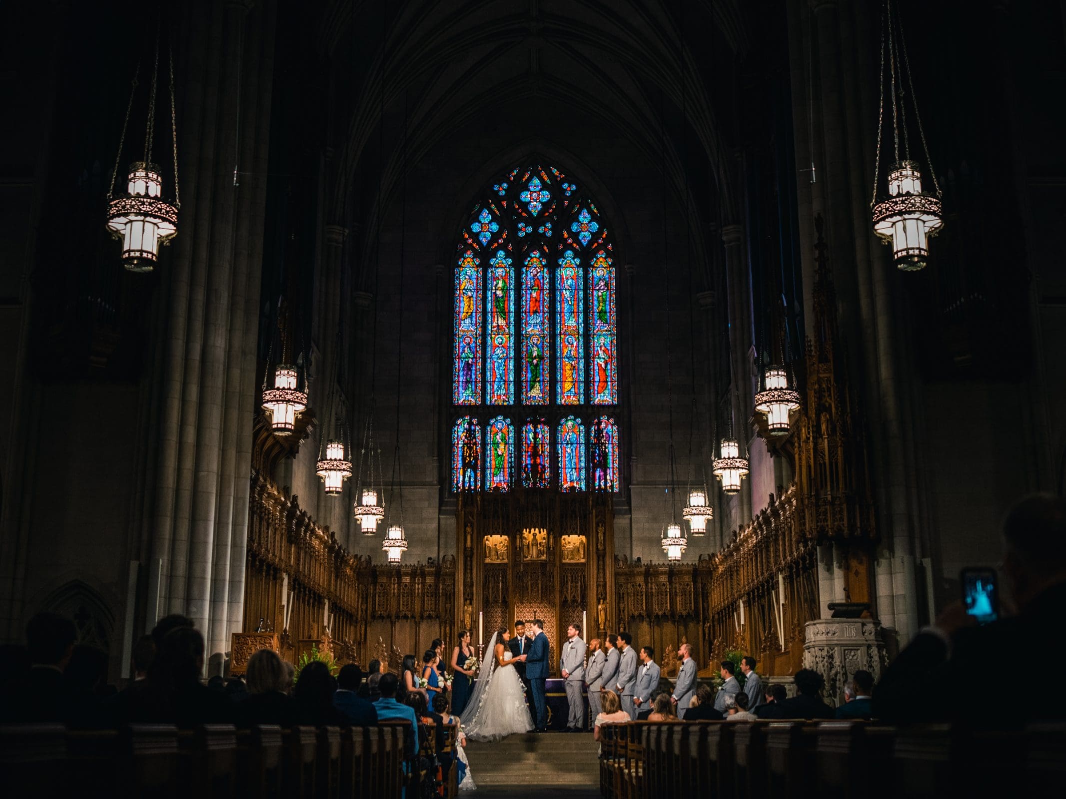 duke chapel & cookery wedding - sasha & ben A wedding ceremony inside the grand Duke Chapel boasts high vaulted ceilings and ornate stained glass windows. The bride and groom stand at the altar surrounded by the wedding party. Guests are seated on wooden pews, with elegant chandeliers casting a warm glow above.