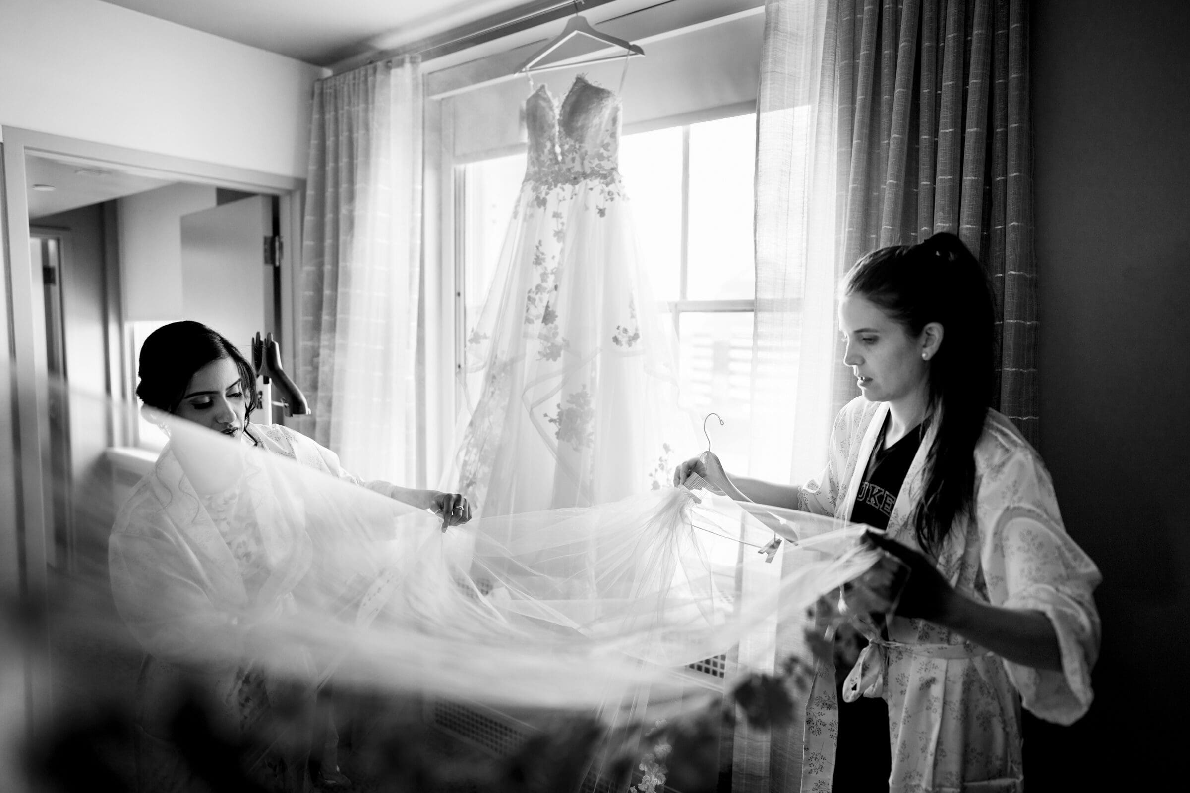 duke chapel & cookery wedding - sasha & ben Two women in a softly lit room hold a delicate veil before a hanging wedding dress, adorned with lace and floral details. The scene evokes serene elegance, reminiscent of Duke Chapel, with natural light filtering through sheer curtains on the window.