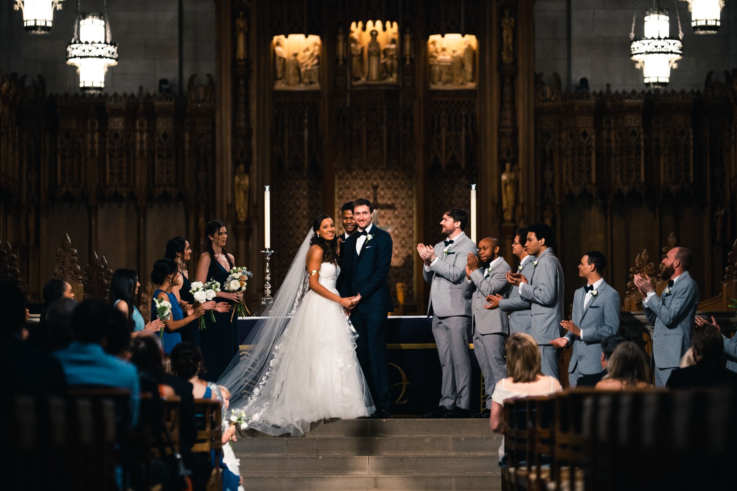 duke chapel & cookery wedding - sasha & ben A bride and groom stand joyfully at the altar in the grandeur of Duke Chapel, surrounded by their bridal party. The bride is in a flowing white gown, while the groom wears a dark suit. The bridal party, dressed in blue and gray, applauds the couple.