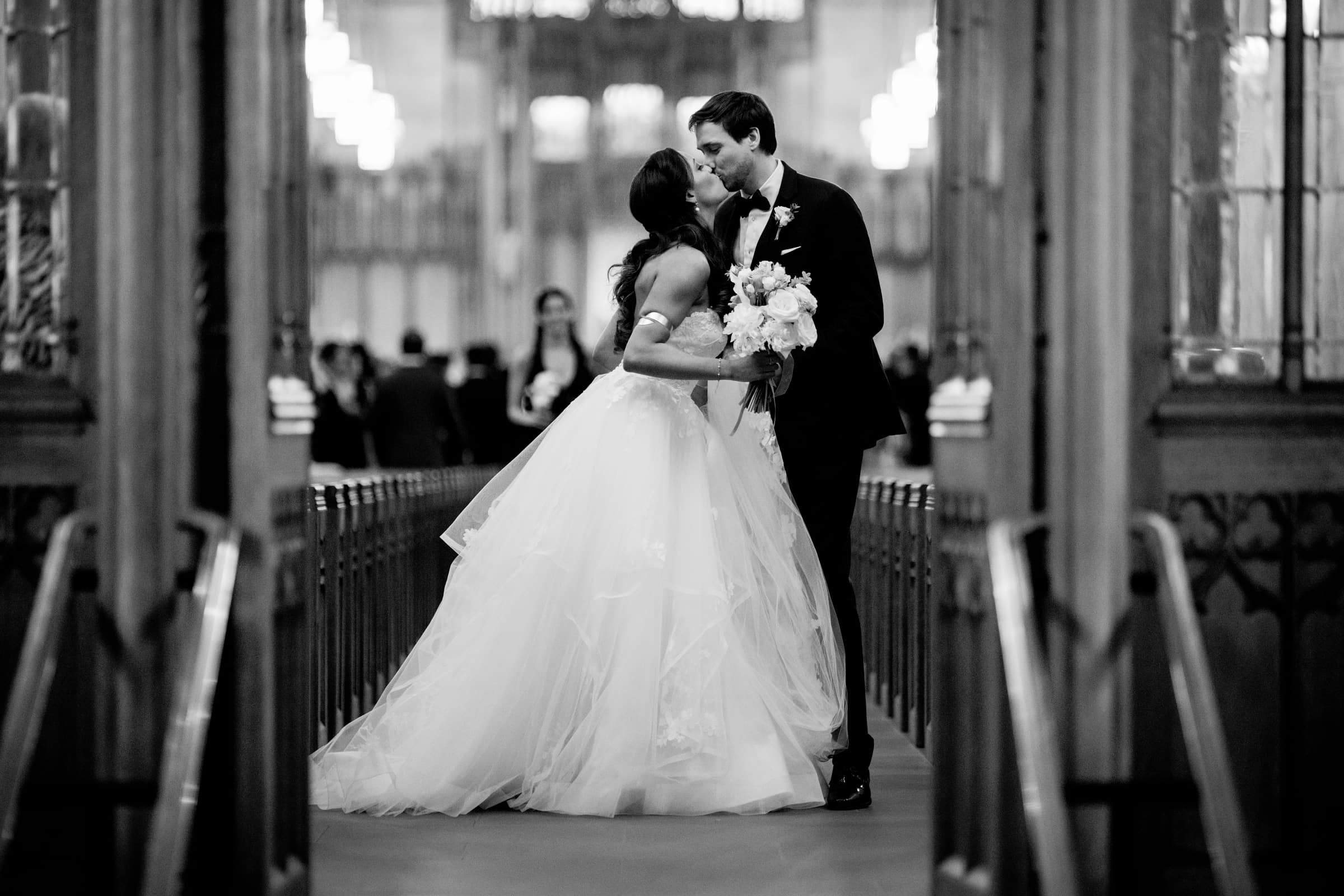 duke chapel & cookery wedding - sasha & ben In the elegant setting of Duke Chapel, a bride and groom share a kiss in the church aisle. The bride, in a flowing white gown, holds a bouquet as the groom stands beside her in his dark suit. The black and white image captures blurred background lights, enhancing the moment's timeless charm.