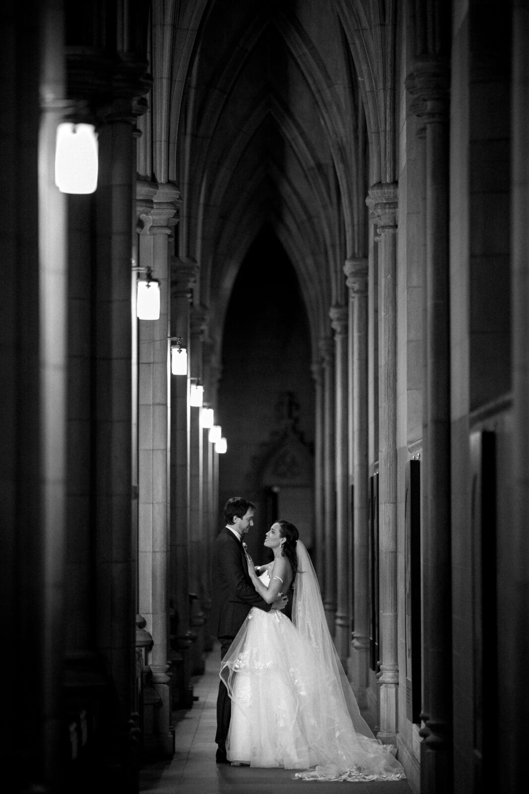 duke chapel & cookery wedding - sasha & ben A black and white photo captures a bride and groom embracing in a cathedral-like corridor reminiscent of Duke Chapel. The arched ceiling and stone columns create a dramatic, romantic atmosphere, as the bride, in her flowing gown and veil, gazes lovingly at her partner.