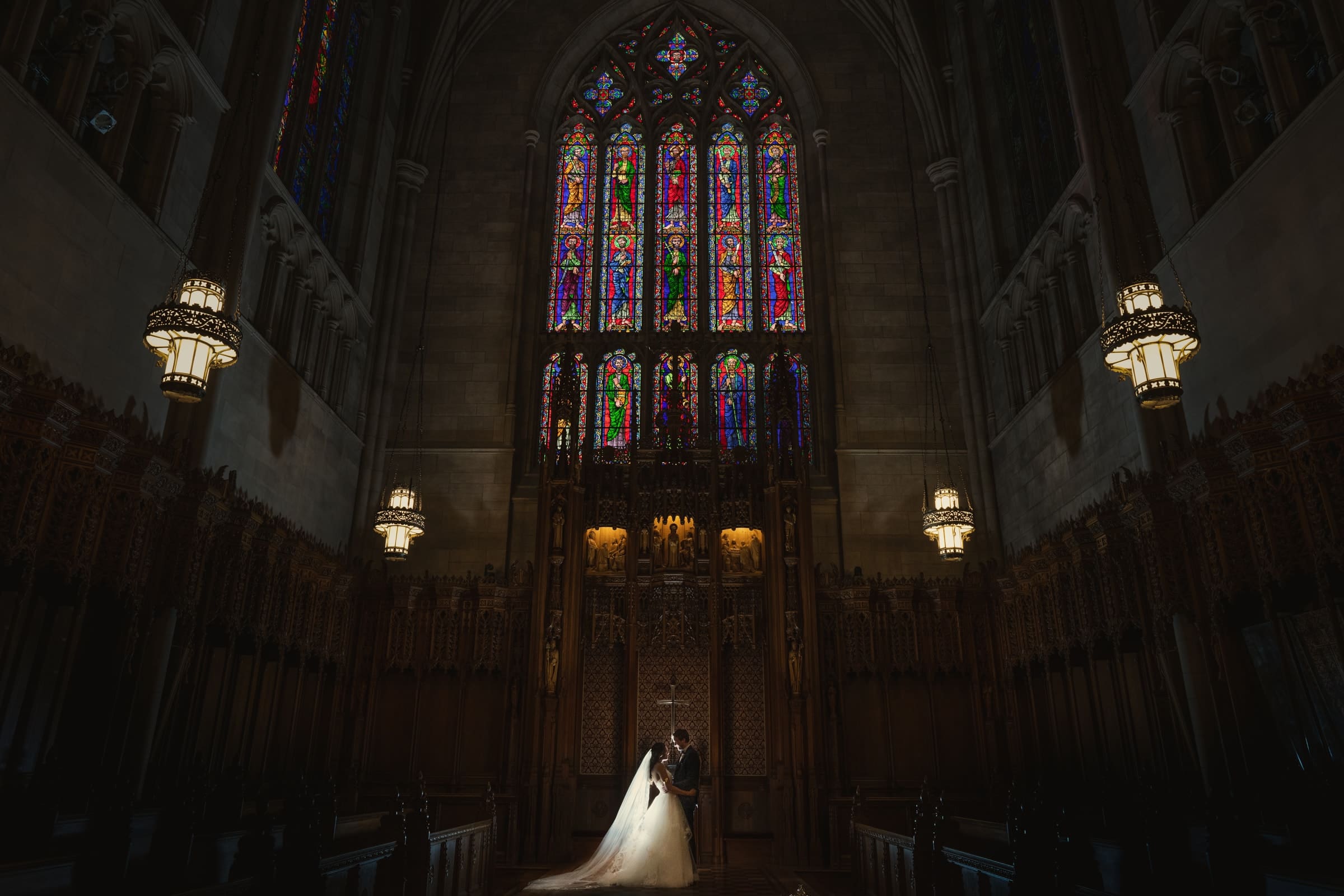 A couple stands at the center of a grand, dimly lit cathedral reminiscent of Duke Chapel. They are illuminated by sunlight streaming through intricate stained glass windows above, creating a colorful backdrop. The arching architecture and hanging lanterns add to the majestic ambiance.