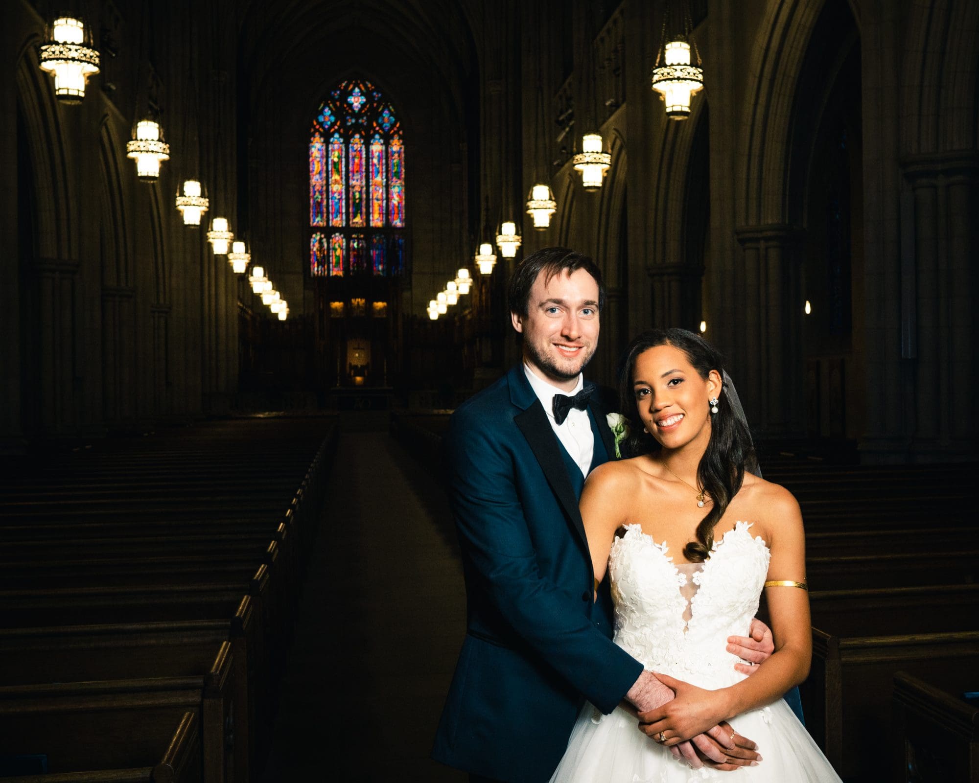 duke chapel & cookery wedding - sasha & ben A couple in formal wedding attire stands in the dimly lit Duke Chapel, with its high arches and stained glass windows. The bride wears a white gown, and the groom is in a black tuxedo, both smiling warmly as soft lighting accentuates the scene.