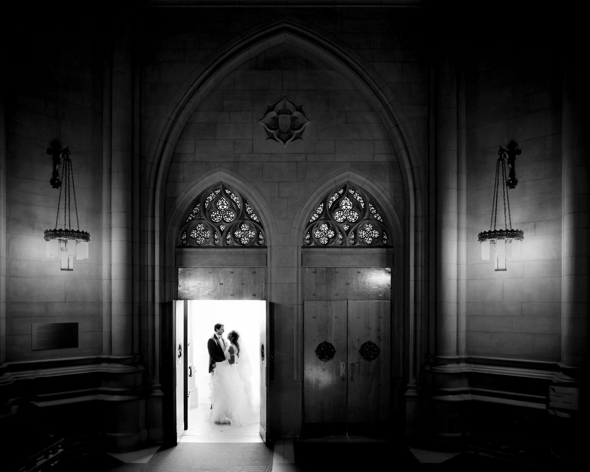 duke chapel & cookery wedding - sasha & ben A bride and groom stand illuminated in a doorway of a dimly lit, gothic-style church reminiscent of Duke Chapel. Ornate wooden doors with decorative carvings frame the couple, while the towering stone walls and arches create a dramatic and romantic atmosphere.