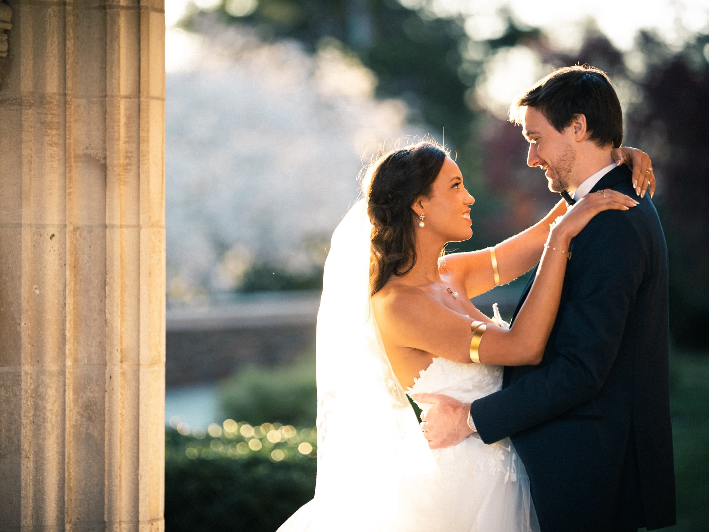 duke chapel & cookery wedding - sasha & ben Outside Duke Chapel, a bride and groom share a tender embrace. The bride, in her white gown and veil, stands with her groom in a dark suit. Sunlight casts a warm glow, highlighting the happy couple against the chapel's stunning backdrop.