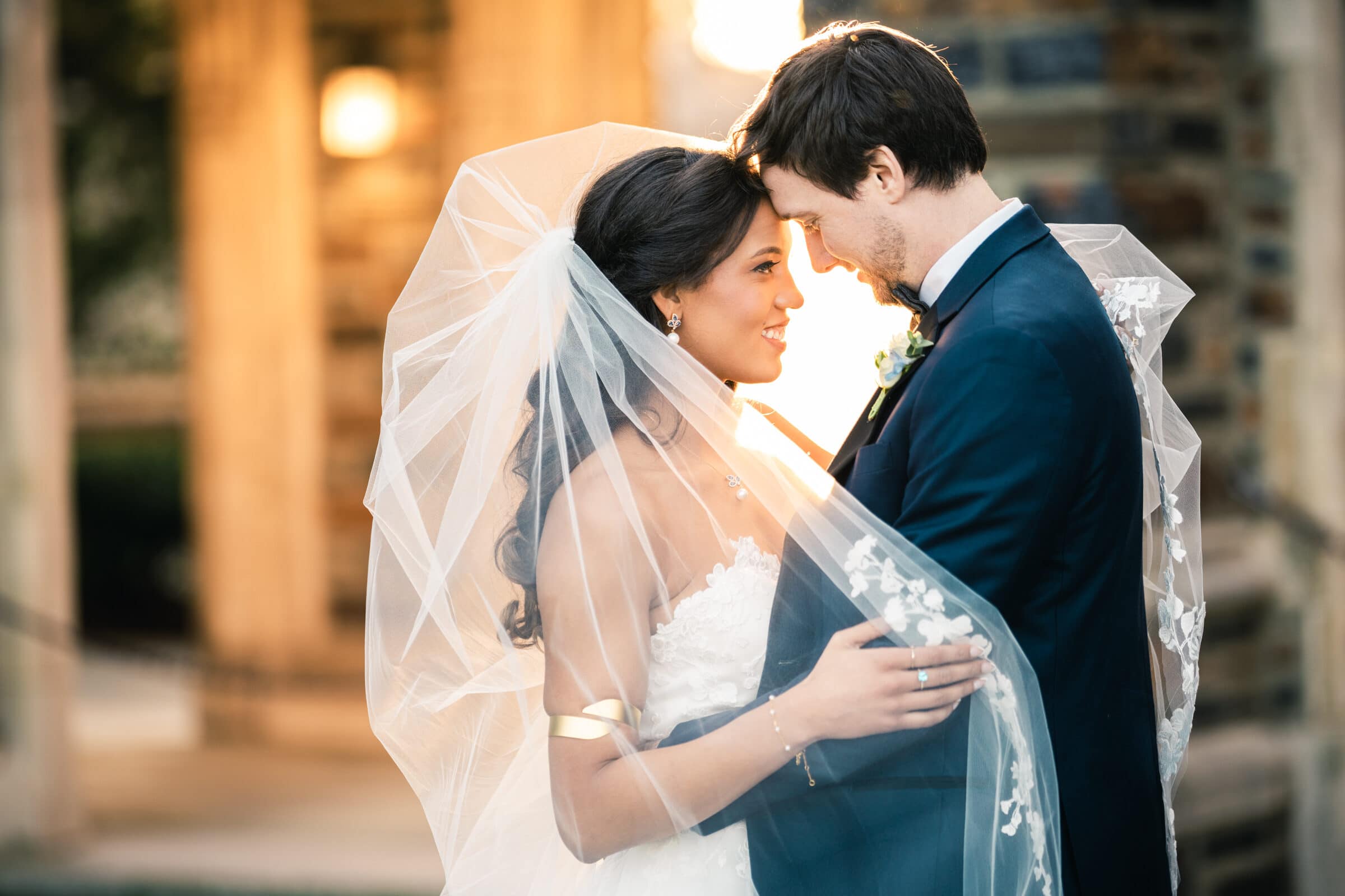 duke chapel & cookery wedding - sasha & ben A bride and groom stand closely, touching foreheads and smiling softly in the warm glow of Duke Chapel. The bride is wearing a white dress with a flowing veil, while the groom is in a black suit, all bathed in romantic ambiance.