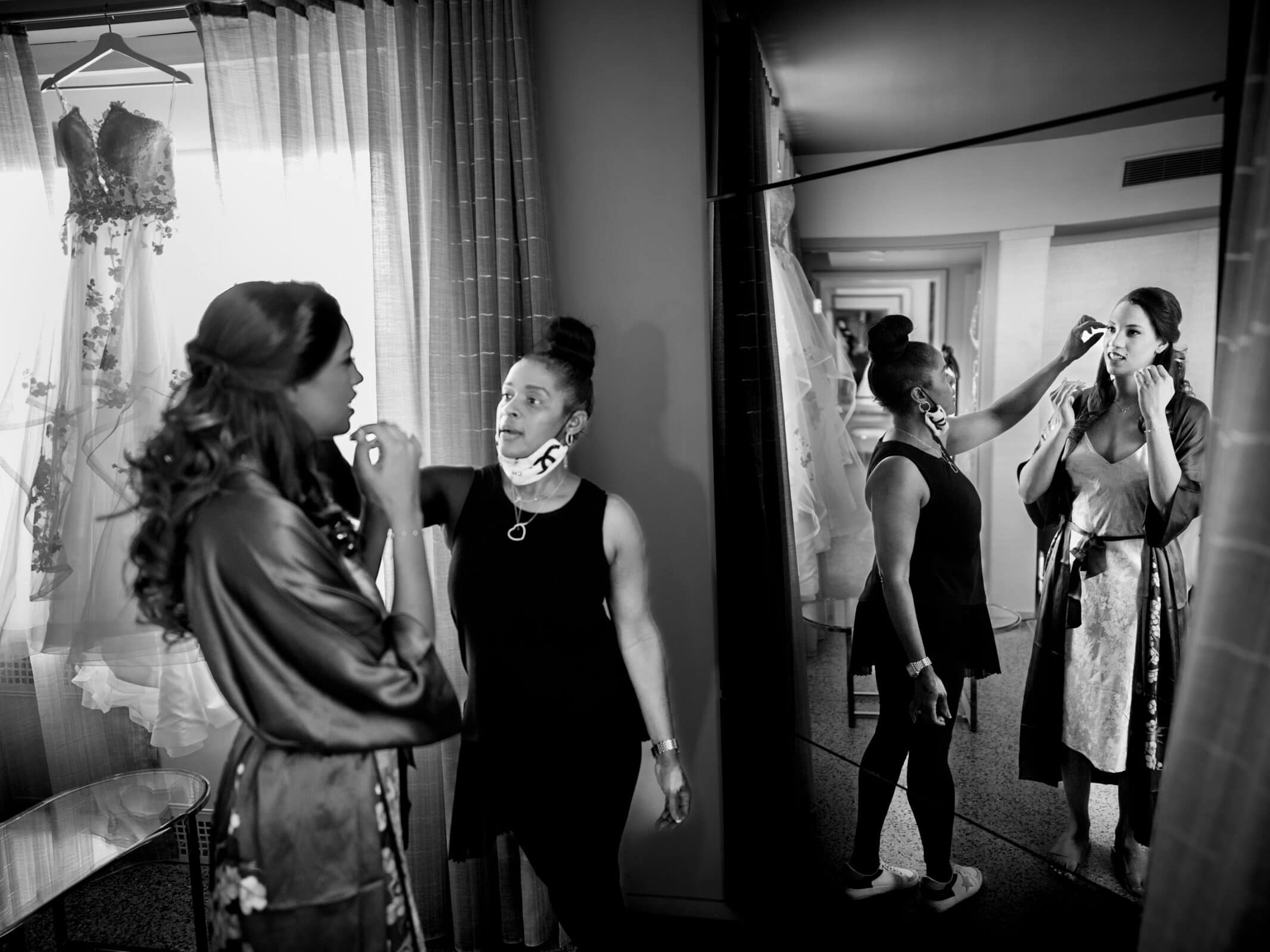 duke chapel & cookery wedding - sasha & ben A woman in a robe getting makeup applied by a stylist stands near a mirrored closet, reflecting their image. A wedding dress hangs nearby, casting a serene ambiance reminiscent of Duke Chapel. The scene is beautifully captured in black and white.