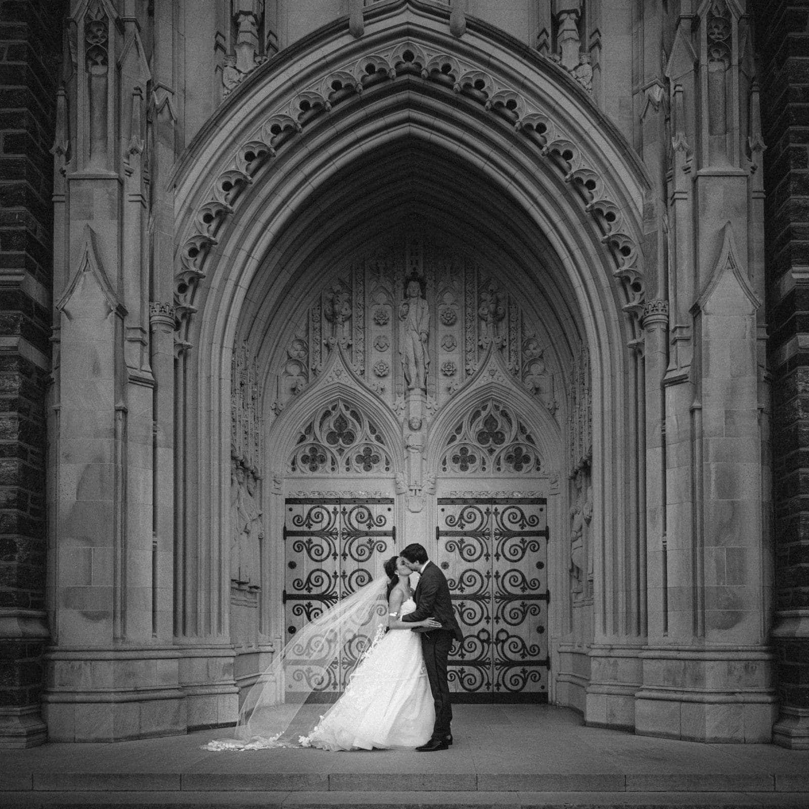 A wedding portrait of a bride and groom embracing under the ornate, carved stone arched entrance of Duke Chapel, with the bride's long veil trailing on the ground by the decorative metal doors.
