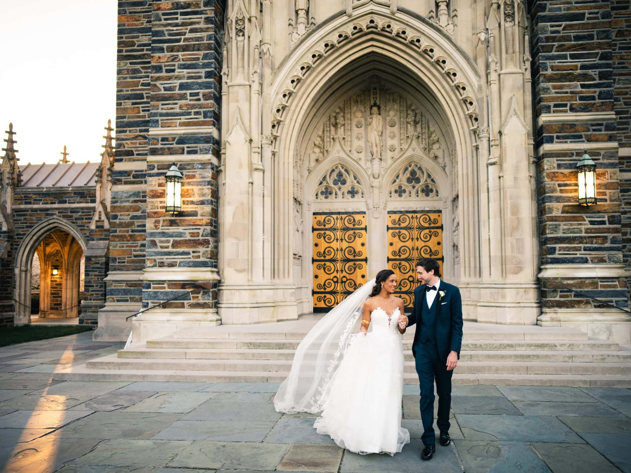 duke chapel & cookery wedding - sasha & ben A bride and groom hold hands, gazing at each other in front of Duke Chapel's ornate, arched entrance with large wooden doors. The bride wears a white gown and veil, while the groom is in a dark suit. The scene is illuminated by soft lighting.