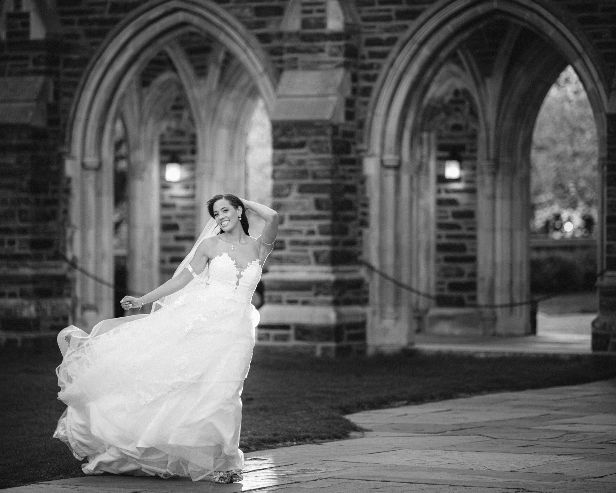duke chapel & cookery wedding - sasha & ben A bride in a flowing white gown twirls joyfully under the stone arches of Duke Chapel. Her long veil catches the wind, and she smiles brightly, creating a sense of movement and happiness against the historic architecture.