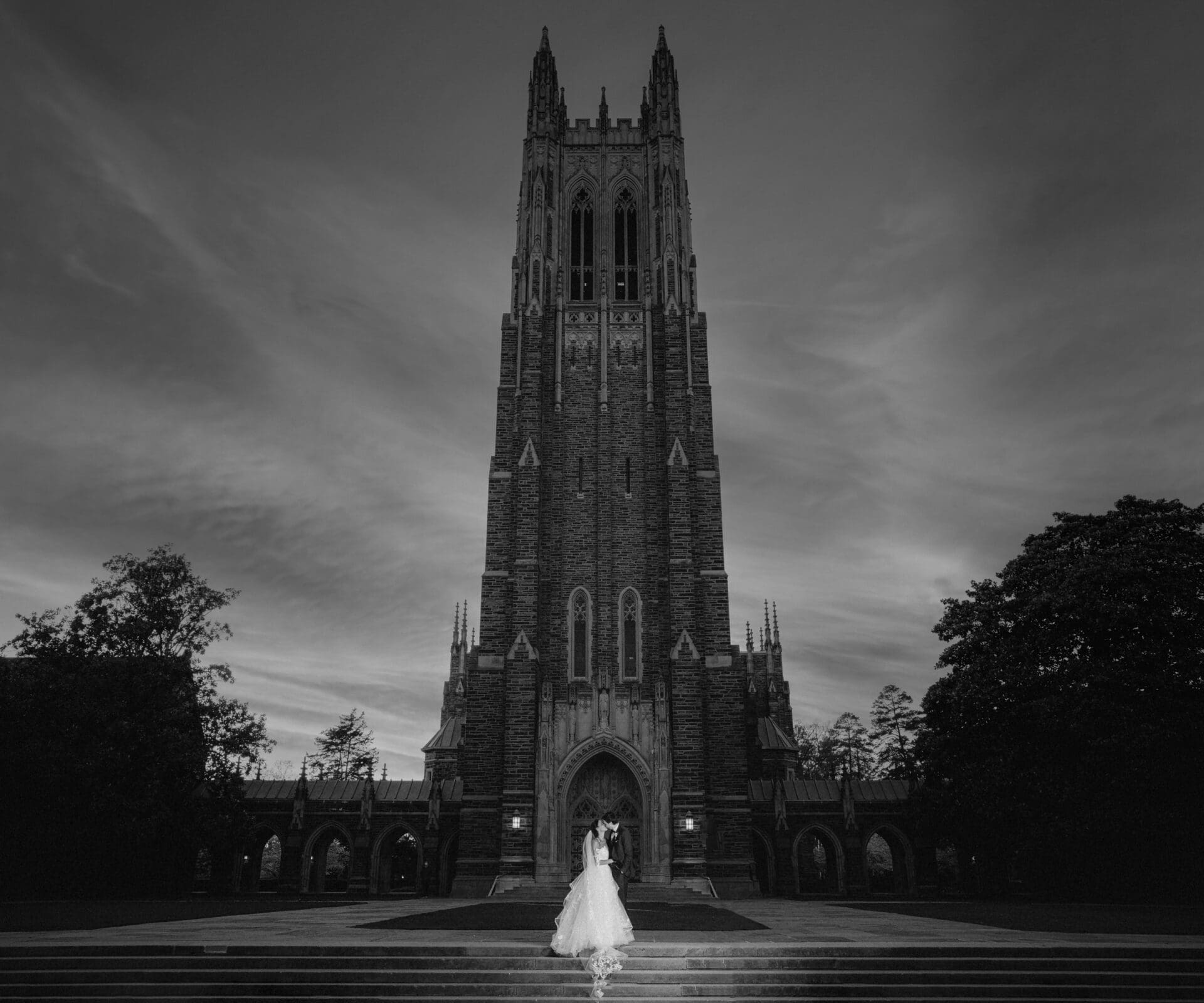 duke chapel & cookery wedding - sasha & ben A couple in wedding attire stands on a wide stone walkway in front of the magnificent Duke Chapel, its spires reaching high under a dramatic cloudy sky. Captured in black and white, the image emphasizes both the chapel's architectural details and the couple's silhouette.