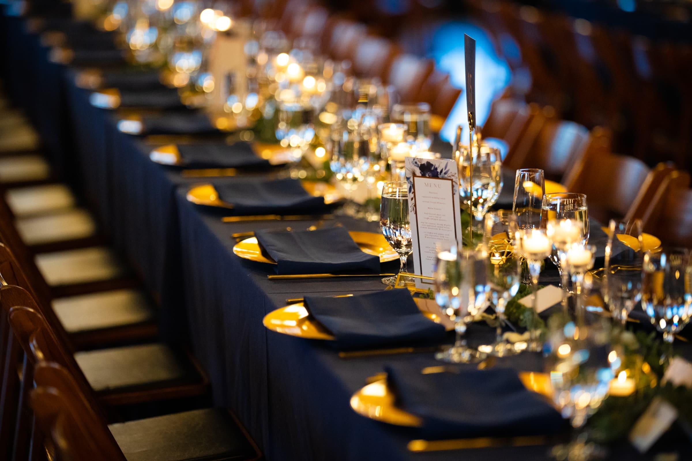 duke chapel & cookery wedding - sasha & ben A long, elegant dining table set for an event at Duke Chapel, featuring navy blue napkins, gold plates, and a menu card at each setting. Glasses of water and wine are placed alongside, with candlelights creating a warm, inviting ambiance.
