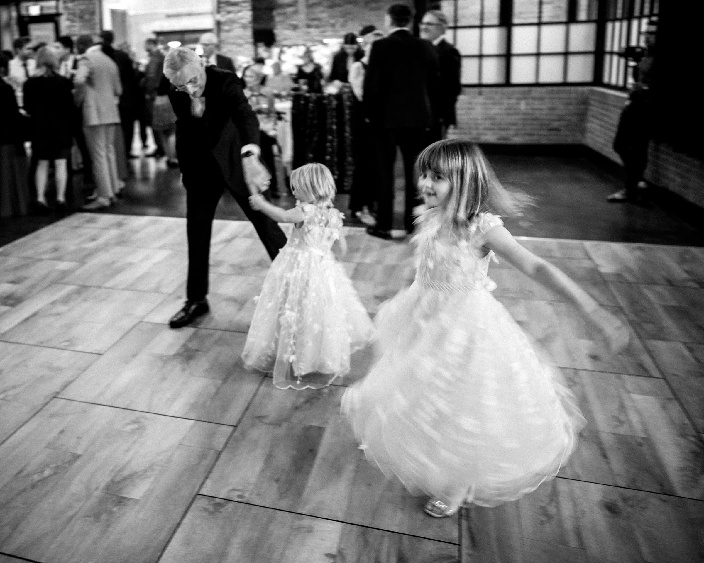 duke chapel & cookery wedding - sasha & ben In a black and white photo, two young girls twirl in dresses on a wooden floor. An adult in a suit, possibly their father, joins one girl. In the background, with a nod to Duke Chapel's grandeur, a group of people socializes in the softly lit room.