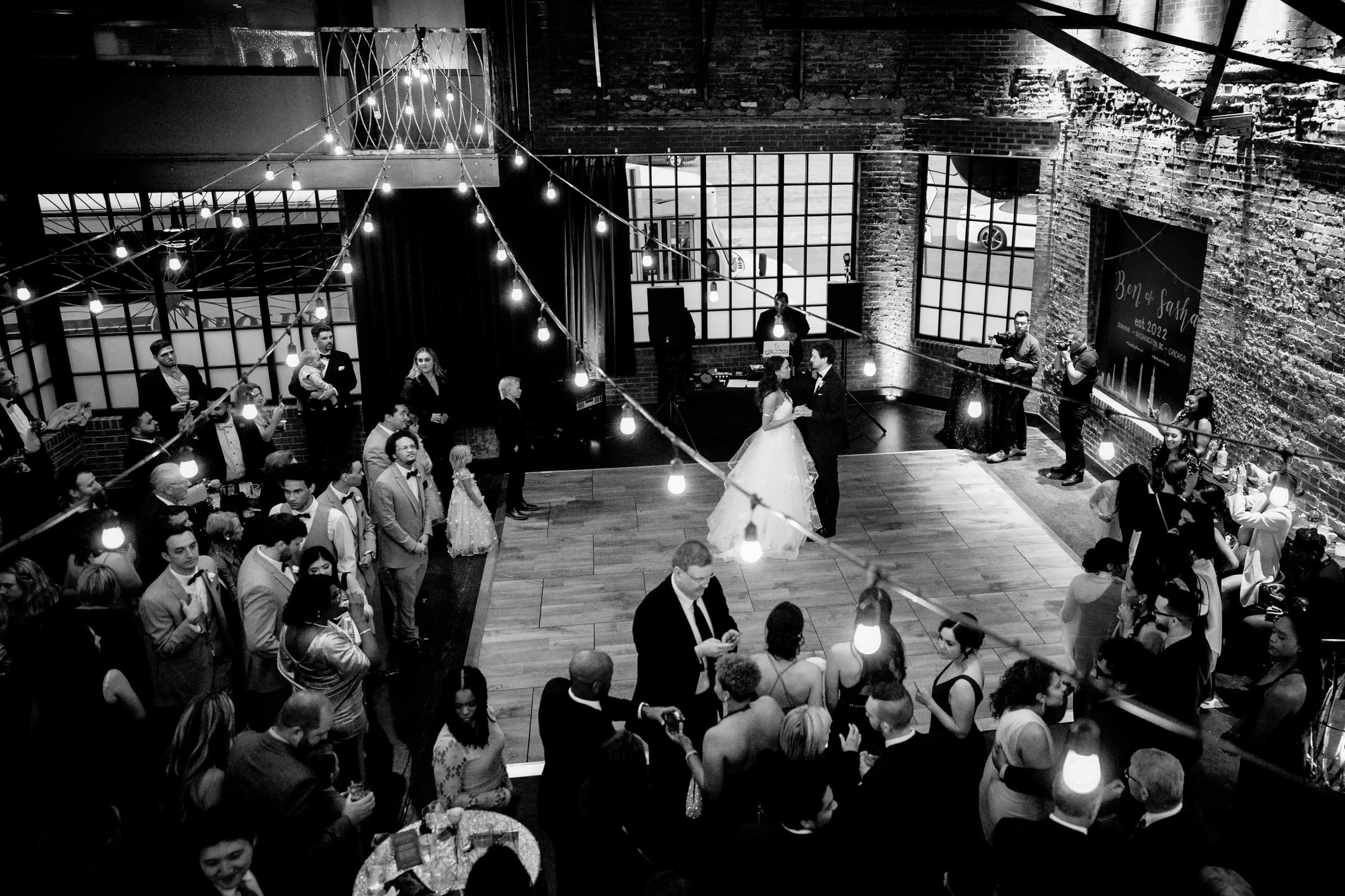 duke chapel & cookery wedding - sasha & ben A black-and-white image captures the magic of a wedding reception in an industrial-style venue reminiscent of Duke Chapel's grandeur. The couple dances in the center as guests surround them on the dance floor, while string lights overhead illuminate the festive atmosphere.