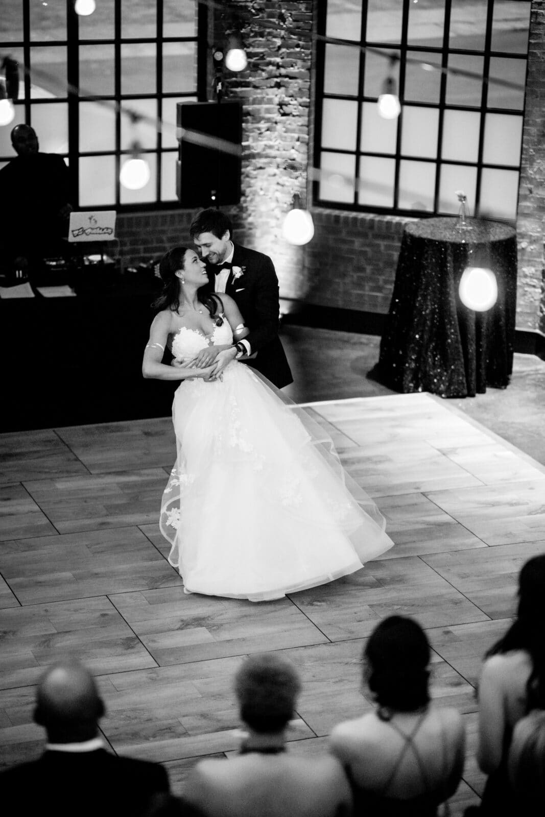 duke chapel & cookery wedding - sasha & ben A bride and groom share their first dance on a wooden floor inside Duke Chapel, softly lit to highlight the romance. The bride wears a flowing white gown, and the groom is in a suit. Guests watch from the foreground as hanging lights and brick walls create an enchanting backdrop.