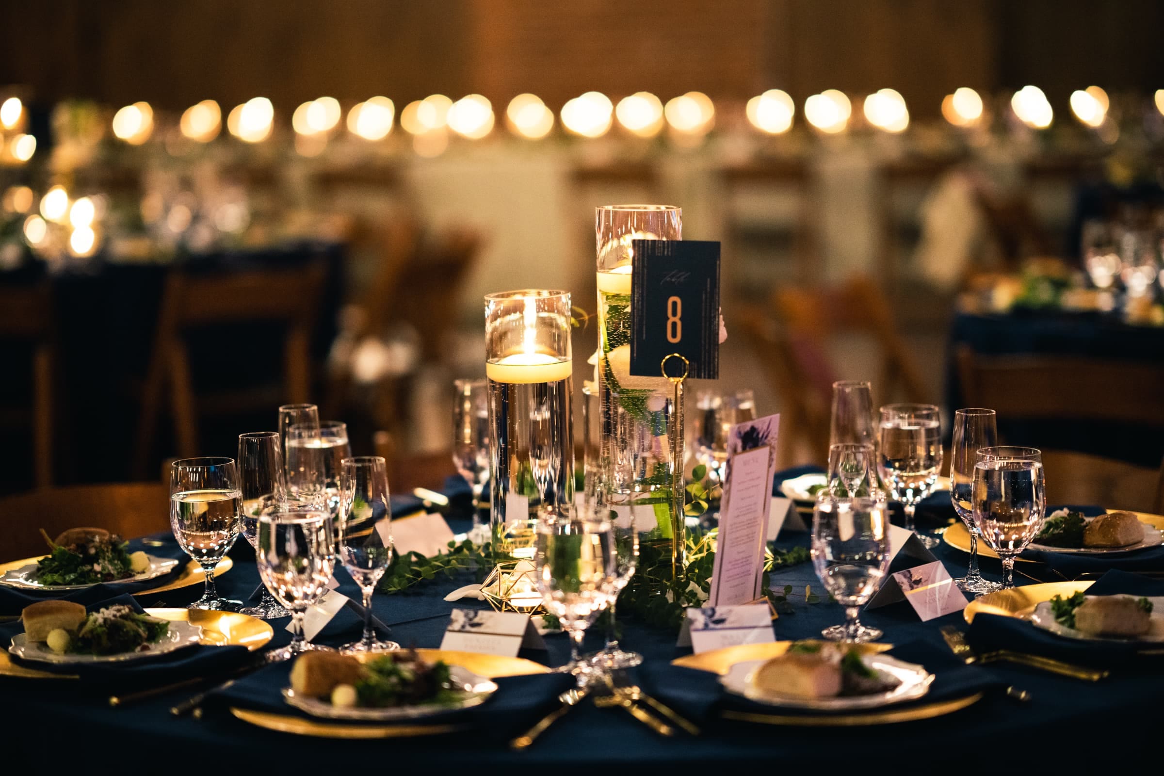 duke chapel & cookery wedding - sasha & ben A beautifully set dining table with elegant glassware, plates, and centerpieces featuring candles in tall holders evokes the grandeur typically found at Duke Chapel events. Plates contain salads and bread rolls on a navy blue tablecloth with a table number 8 card. The background is softly lit.