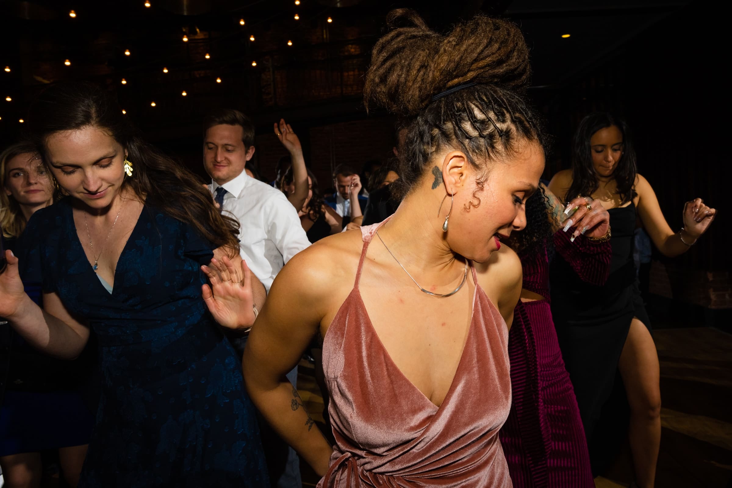duke chapel & cookery wedding - sasha & ben A group of people dancing energetically in a dimly lit room reminiscent of Duke Chapel. The focus is on a woman with braided hair wearing a pink dress. Other dancers, dressed in formal attire, are visible in the background. String lights create a warm atmosphere.