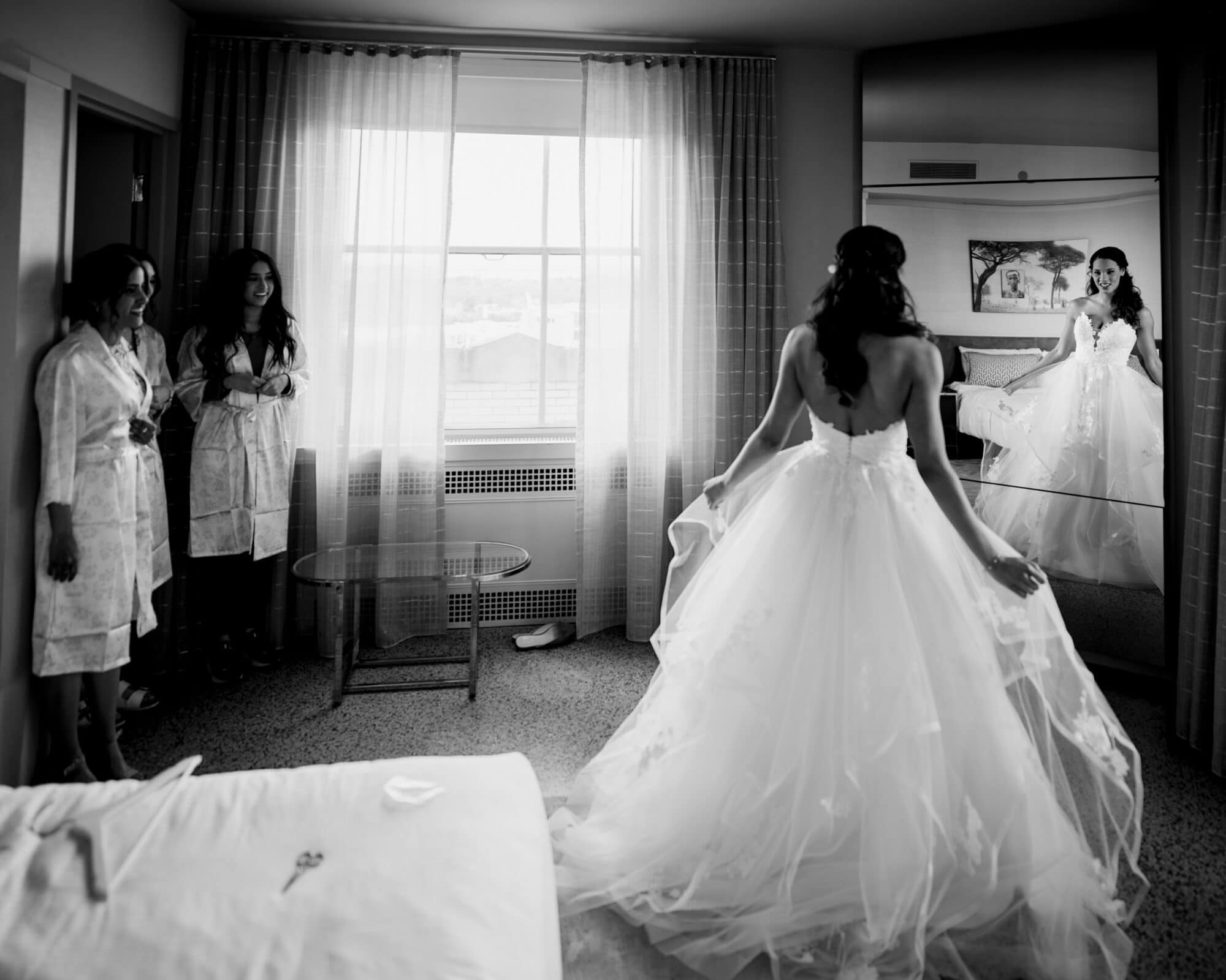 duke chapel & cookery wedding - sasha & ben A bride in a flowing gown stands in front of a mirror in a hotel room, adjusting her dress. Two women in robes look on by the window, which lets in natural light. The scene captures a calm, intimate pre-wedding moment, reminiscent of those cherished Duke Chapel weddings.