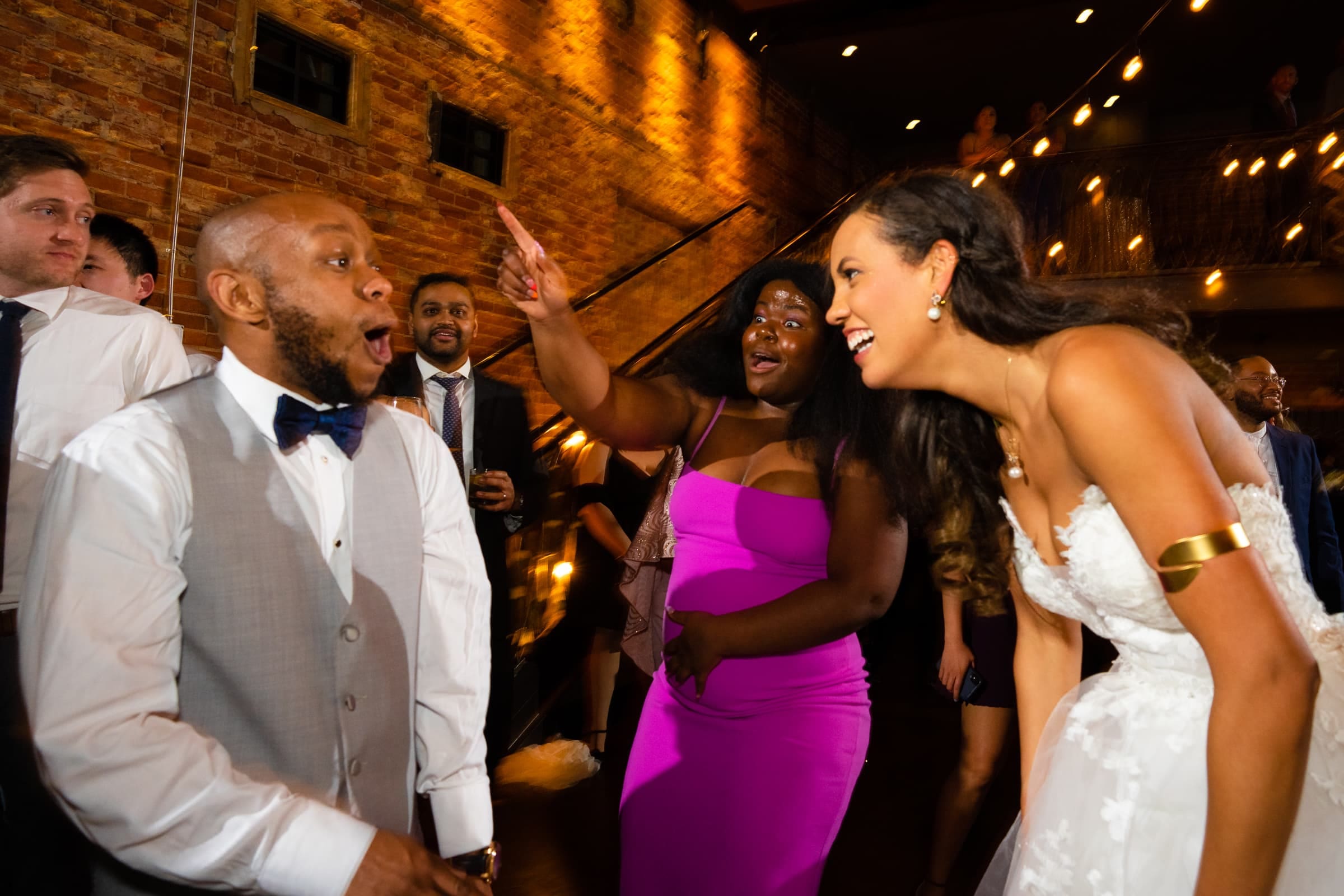 duke chapel & cookery wedding - sasha & ben A joyful wedding scene unfolds at Duke Chapel, with a bride in a white dress laughing with guests. A man in a vest and bow tie appears surprised, while a woman in a pink dress points playfully. The festive atmosphere is highlighted by warm lighting and a brick wall backdrop.