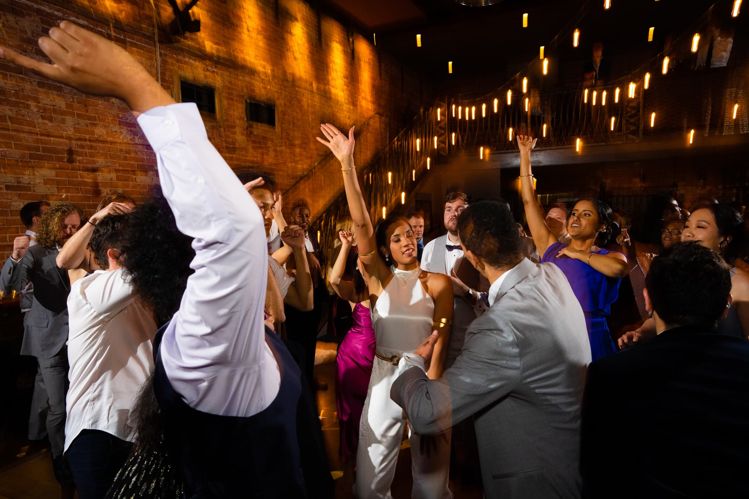 duke chapel & cookery wedding - sasha & ben People are dancing energetically at a lively indoor event near Duke Chapel. The atmosphere is joyful, with individuals raising their hands and enjoying the music. The venue has warm, ambient lighting with strings of lights in the background, enhancing the festive mood.