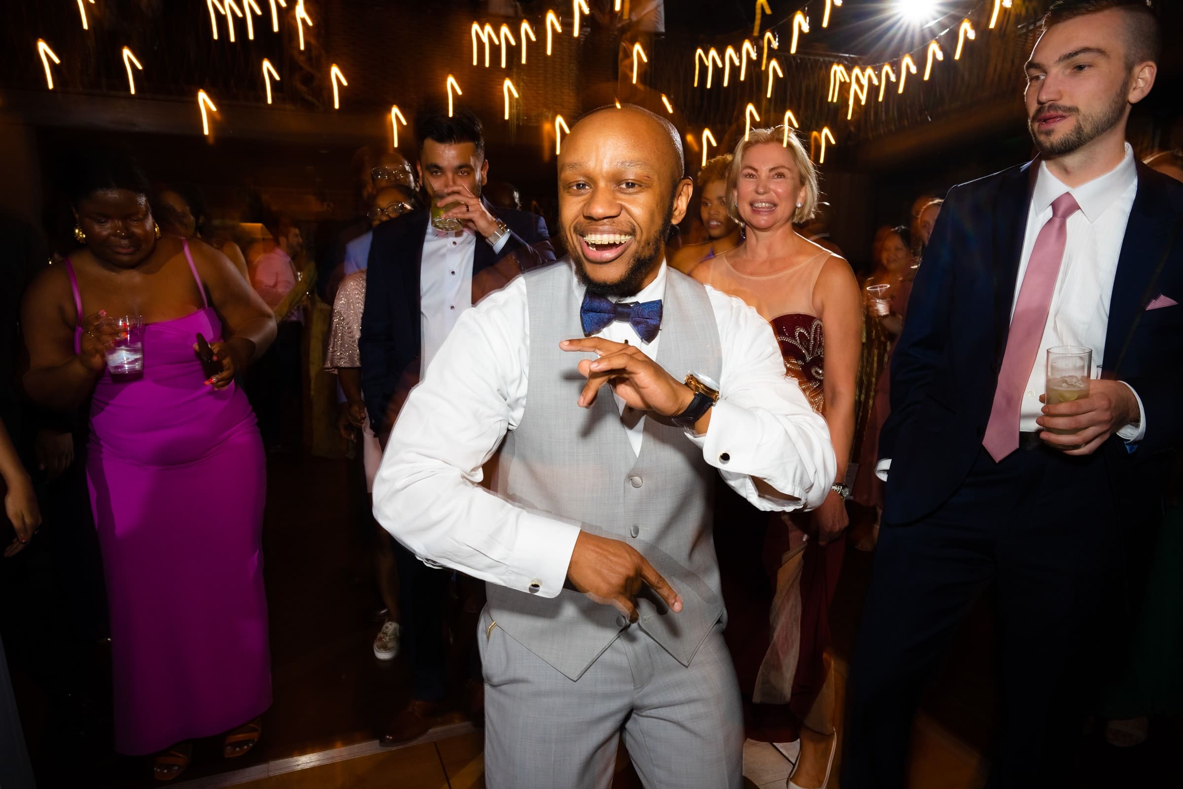 duke chapel & cookery wedding - sasha & ben A joyful man in a light gray suit and bow tie dances enthusiastically at a lively party near Duke Chapel. Several people in formal attire surround him, smiling and holding drinks, with festive string lights illuminating the background.