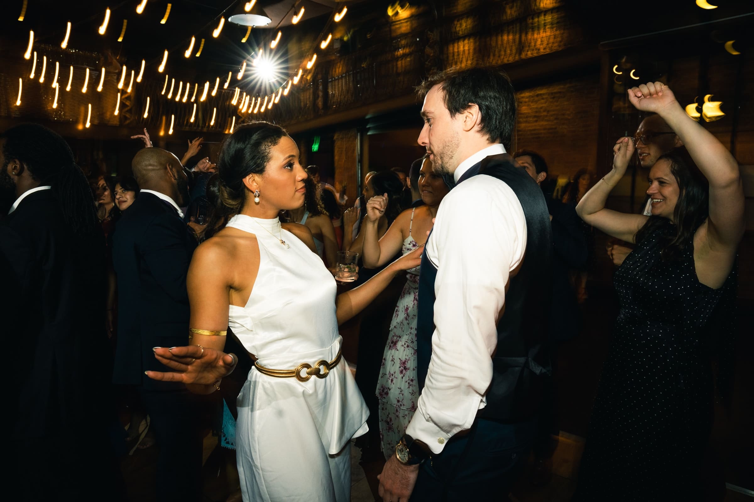 duke chapel & cookery wedding - sasha & ben A lively wedding reception at Duke Chapel features a couple dancing under warm string lights. The woman wears a white dress, and the man is in a vest and tie. Guests around them are enjoying the celebration.